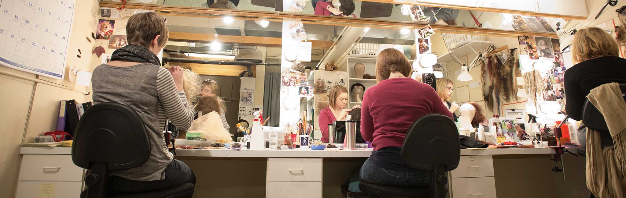 Three people sitting in chairs facing a large mirror working on wigs.