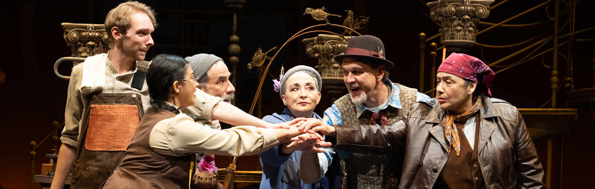 Cast members on a theater stage gather in a circle, reaching hands together around a blue suitcase.