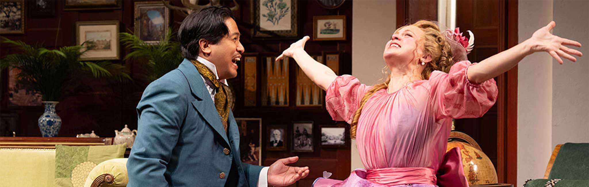 Two people in period costumes in a decorated room; one kneels, the other sits with raised arms.