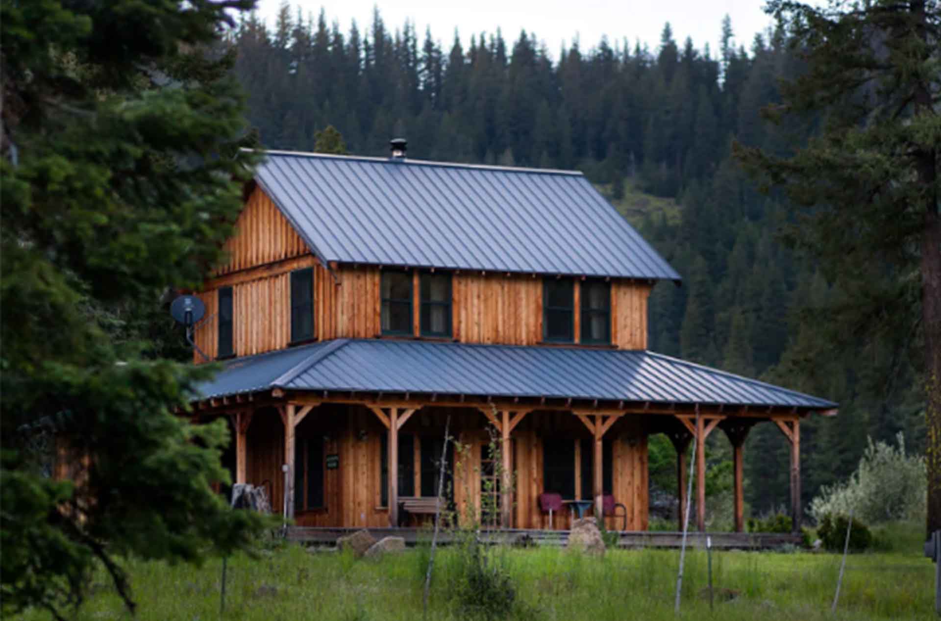Rustic two-story wooden cabin with a metal roof, surrounded by lush greenery and dense forest trees in the background. The cabin features a wide porch with seating, blending into a peaceful natural landscape.
