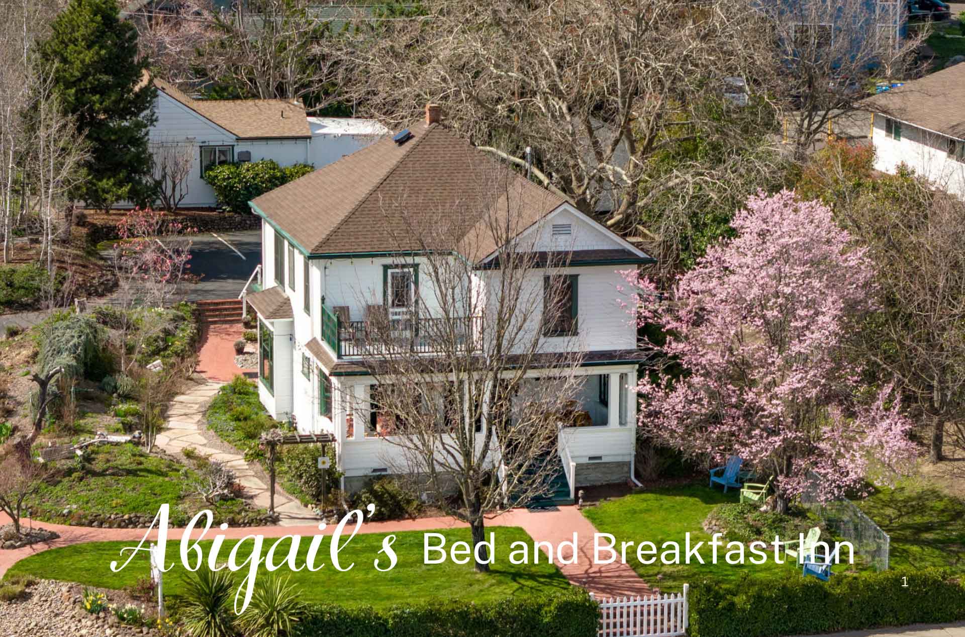 Aerial view of a bed and breakfast inn with a white house, a blooming pink tree, a brick pathway, and well-maintained gardens. The text reads "Abigail's Bed and Breakfast Inn.