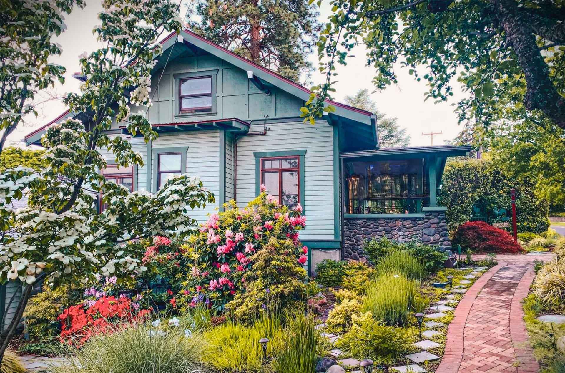 A charming green craftsman-style house surrounded by lush landscaping with vibrant flowers, a stone pathway, and trees framing the scene.