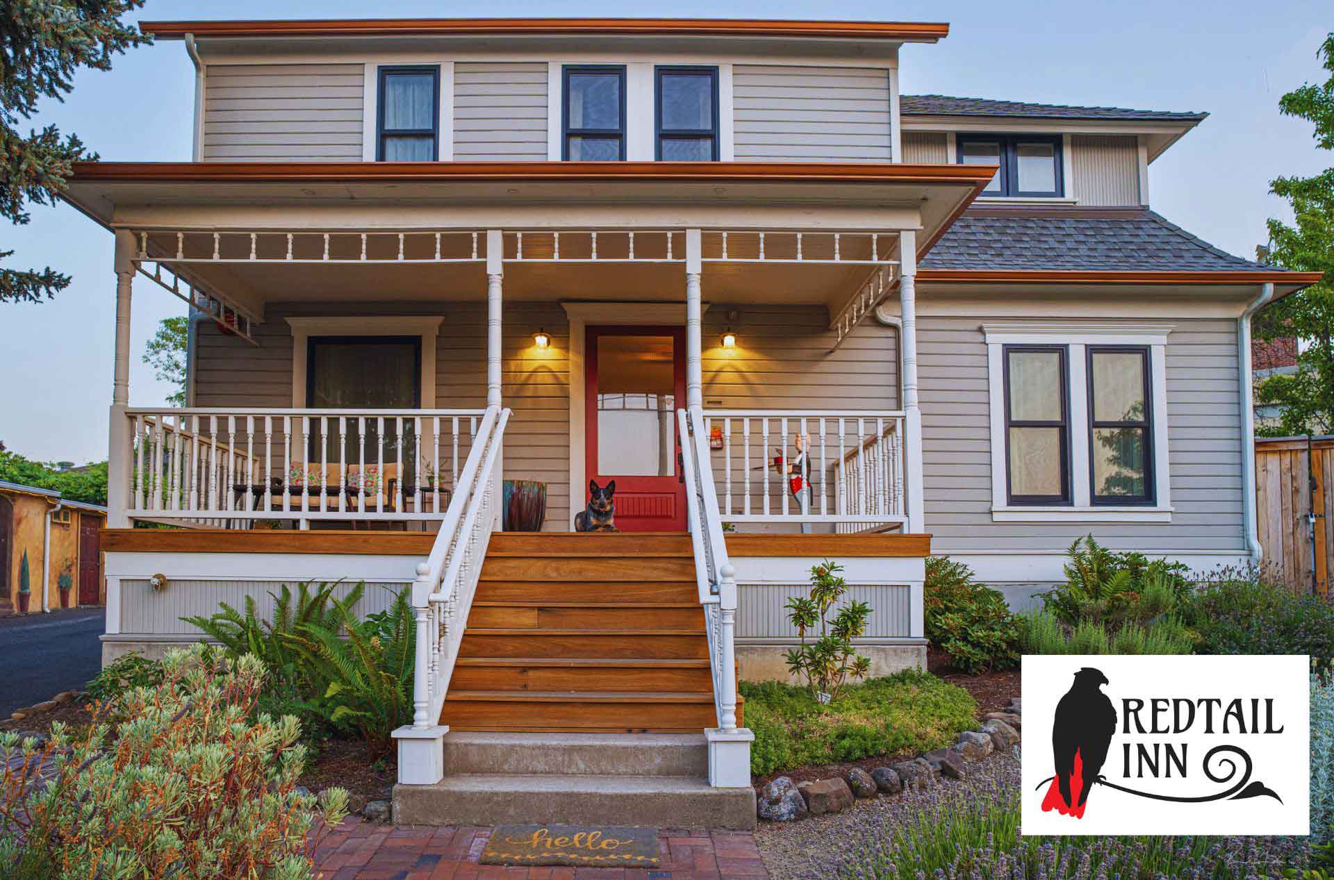 A cozy two-story white house with a welcoming front porch, a wooden staircase, and a dog sitting near the front door, accompanied by the Redtail Inn logo in the bottom corner.