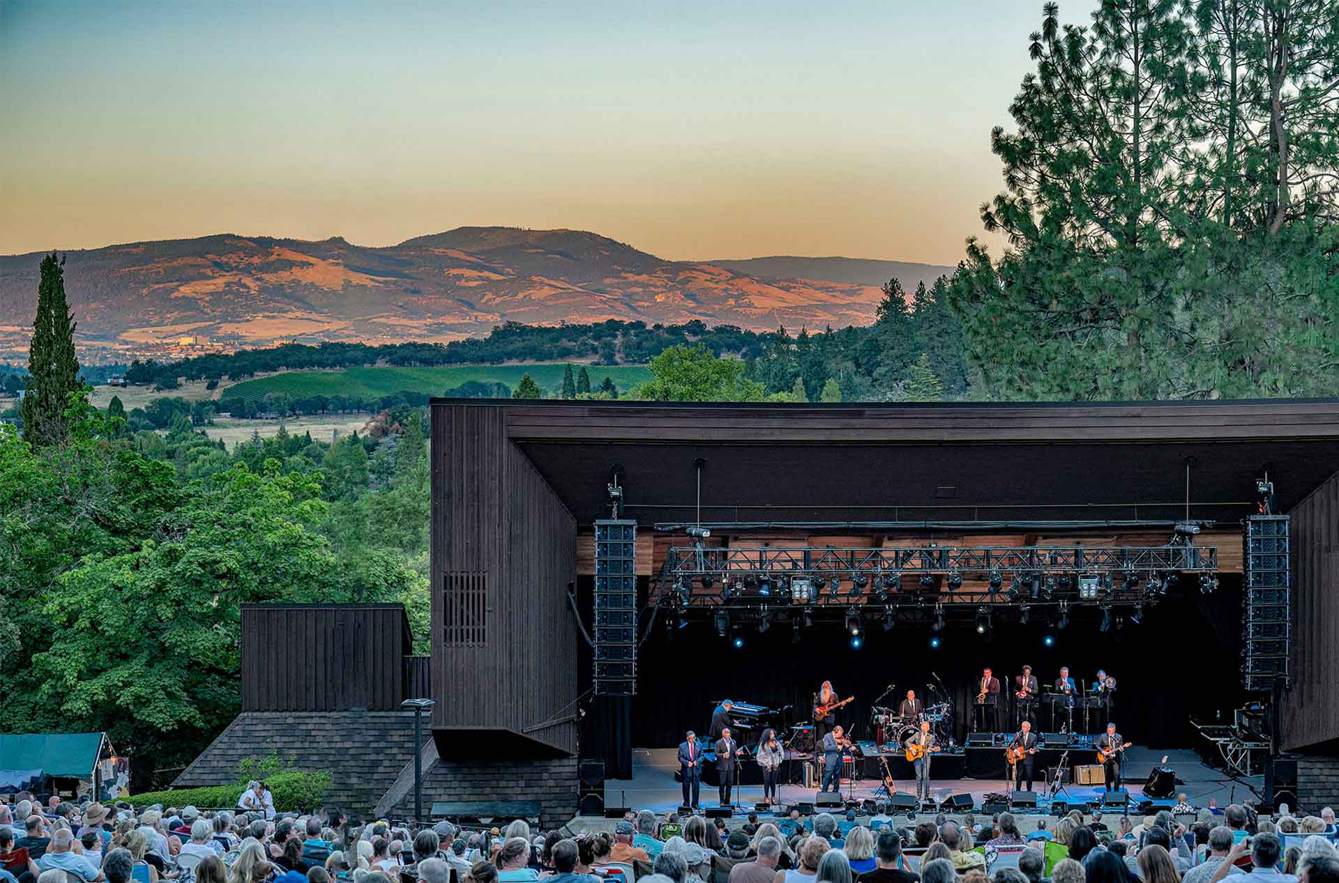 Outdoor concert at a hillside amphitheater with a band performing on stage in front of a seated crowd. Rolling green vineyards and sunlit hills stretch into the distance behind the venue under a clear evening sky.