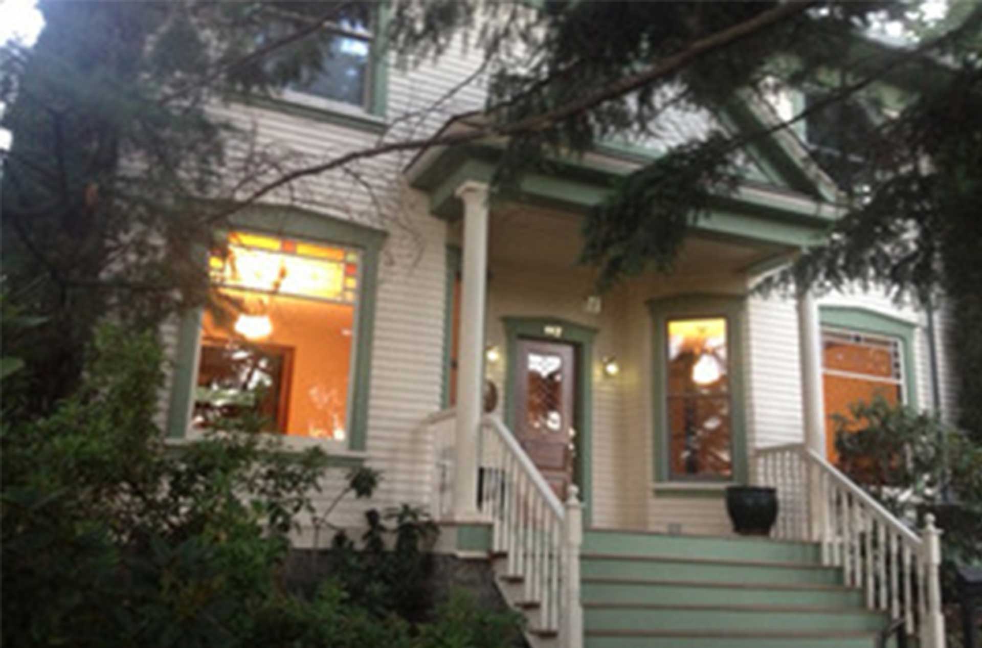 White house with green trim and a lit front porch partially hidden by tree branches.