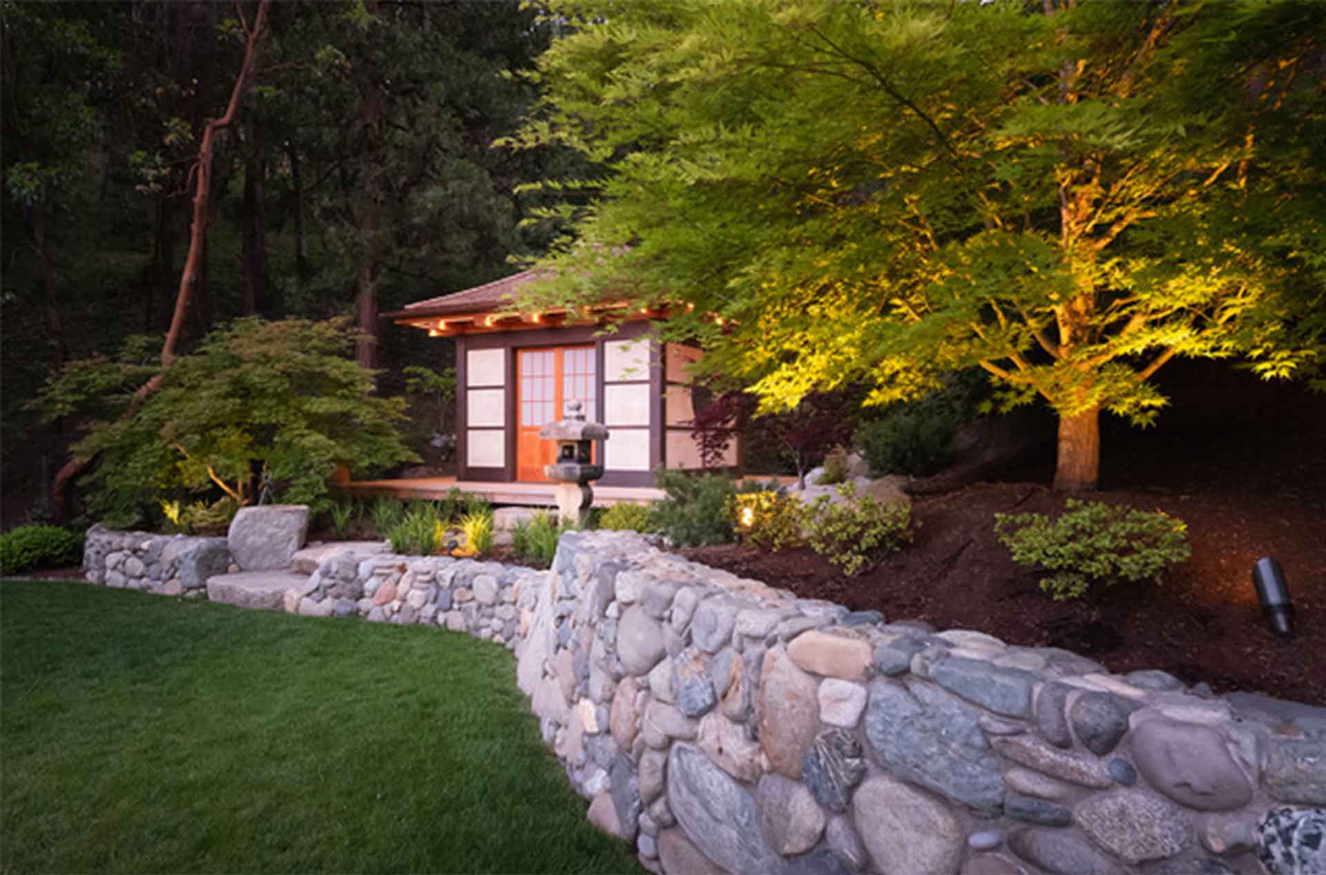 A small Japanese style garden pavilion sits among lush trees and shrubs, softly lit at dusk. A curved stone retaining wall borders a manicured lawn in the foreground, creating a calm, landscaped setting.