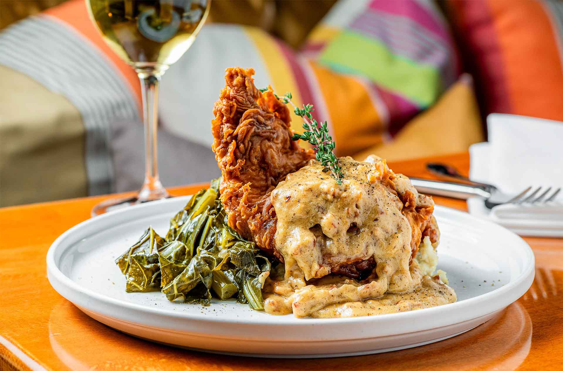 Plate of crispy fried chicken topped with creamy gravy, served with collard greens and mashed potatoes, with a glass of white wine in the background.