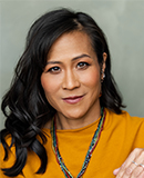 Professional headshot of a person with long dark hair wearing a mustard-yellow top and a multicolored necklace, facing the camera against a neutral gray background. 