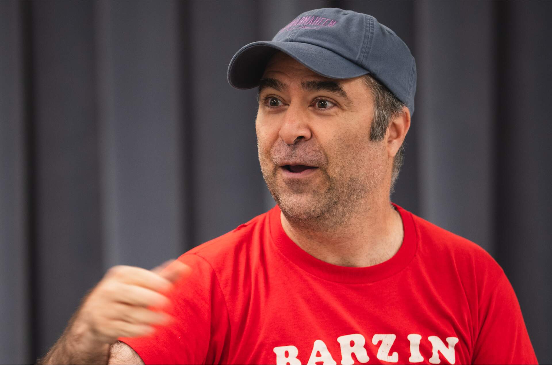 A  person wearing a red t-shirt with "BARZIN" printed on it and a dark blue baseball cap is sitting on a chair at a table. They seem engaged in conversation, gesturing with their right hand. The background features a curtain.