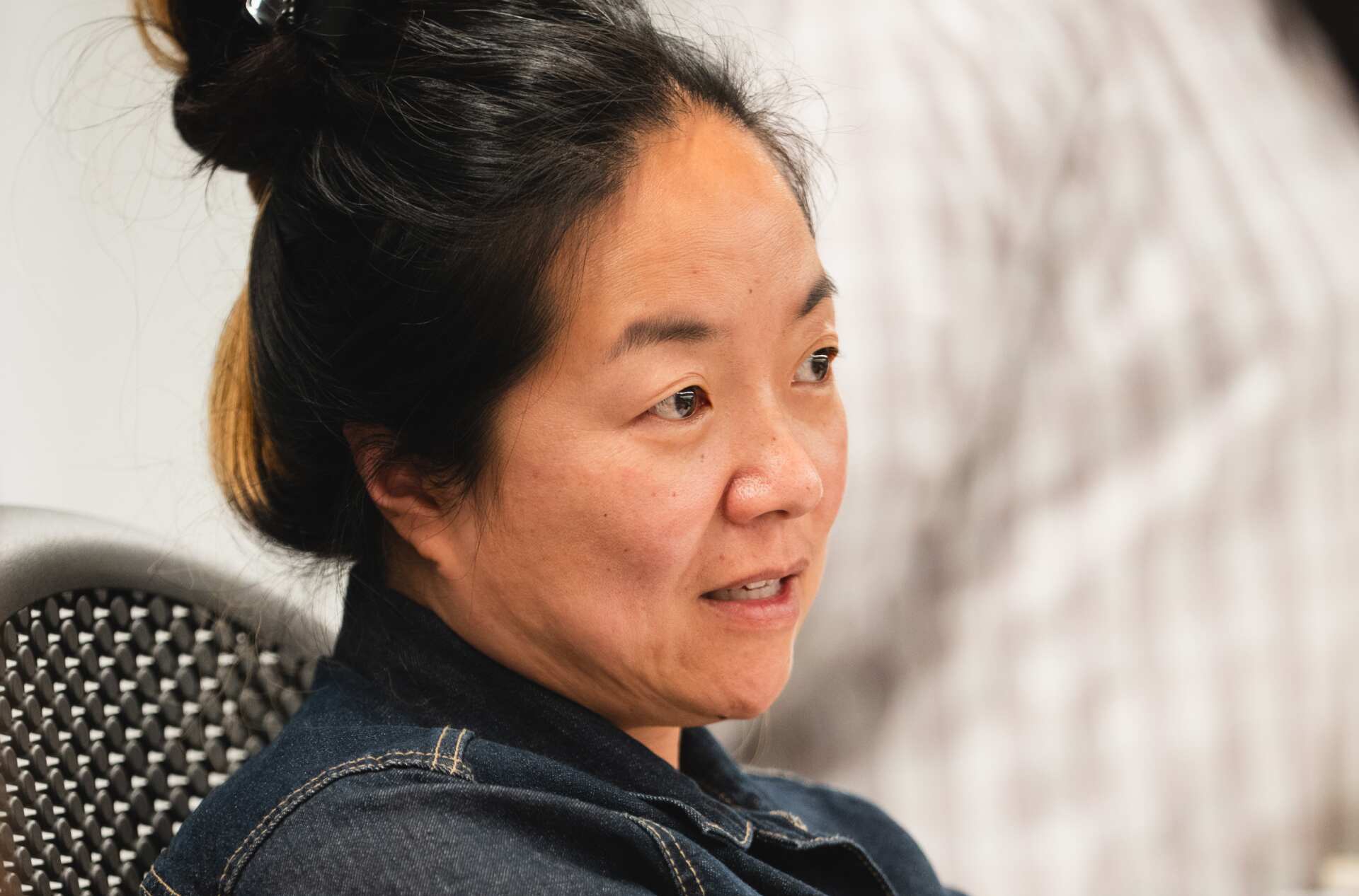 A person with hair tied up is seated on an office chair, wearing a dark jacket, and has arms crossed.