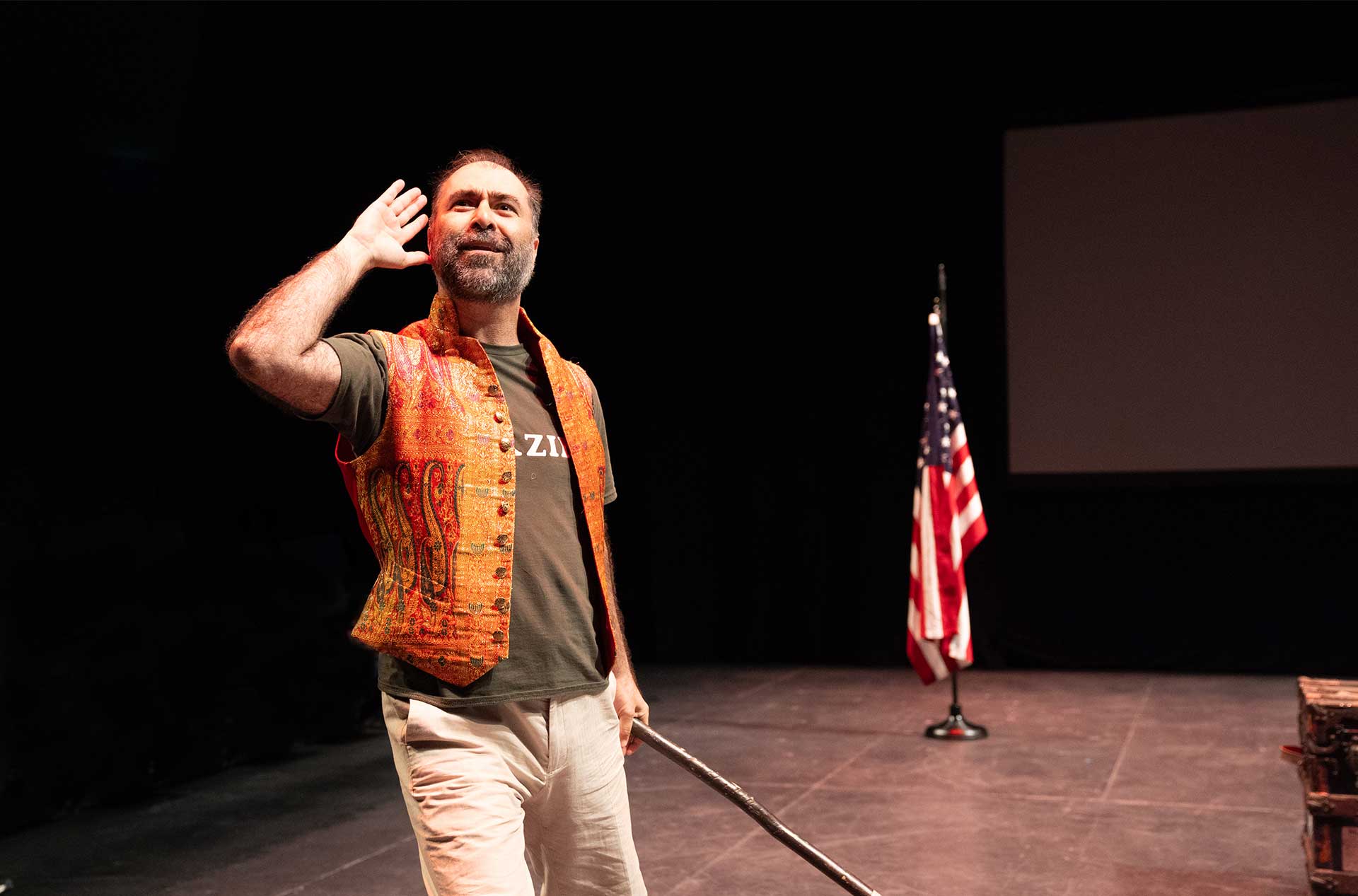 A person cups a hand to their ear as if listening, holding a stick, while wearing an orange vest on stage with an American flag in the background.
