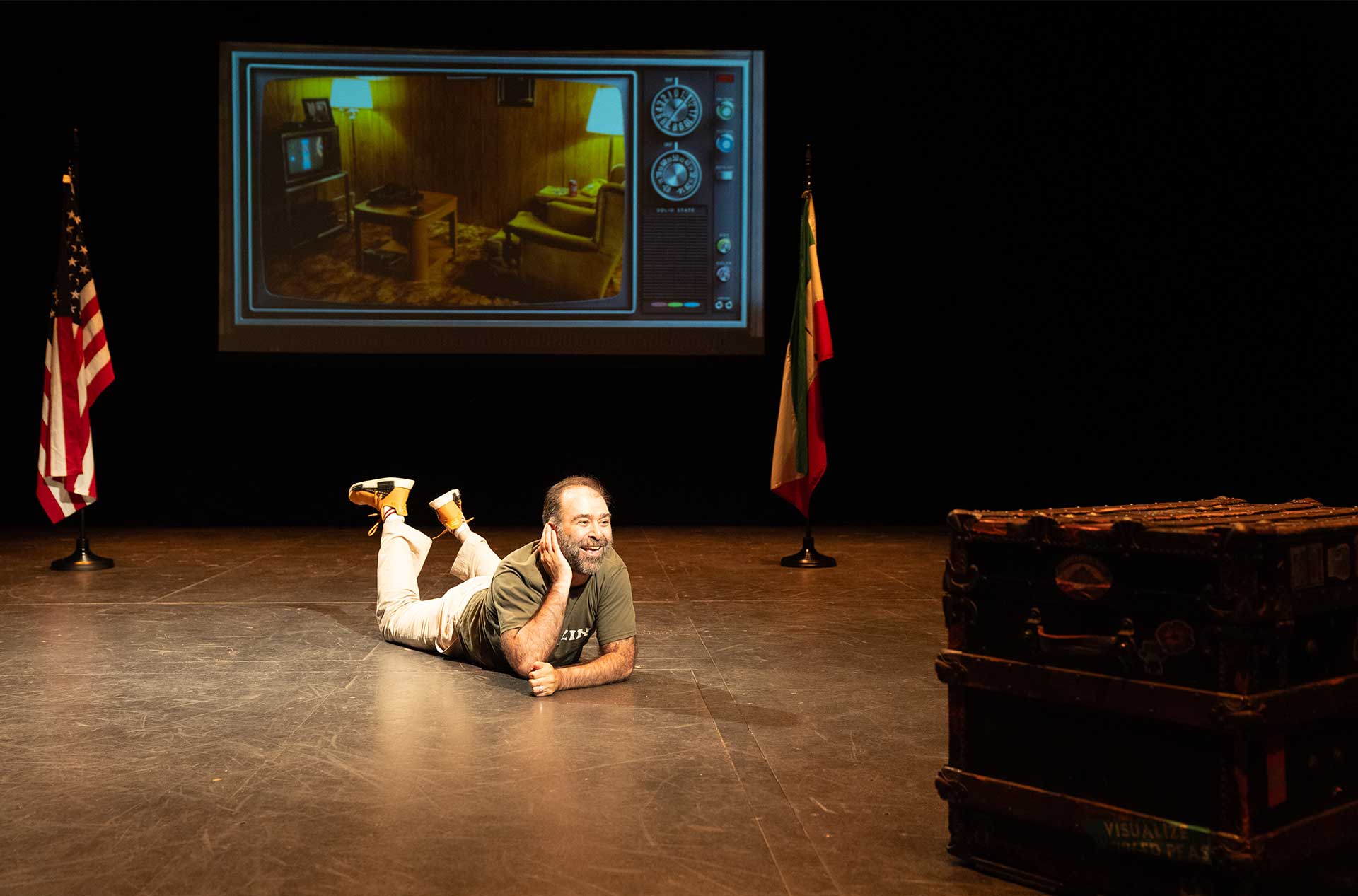A person lies on the stage floor with a cheerful expression, propping up their head with one hand, while a screen behind them shows a vintage living room scene.