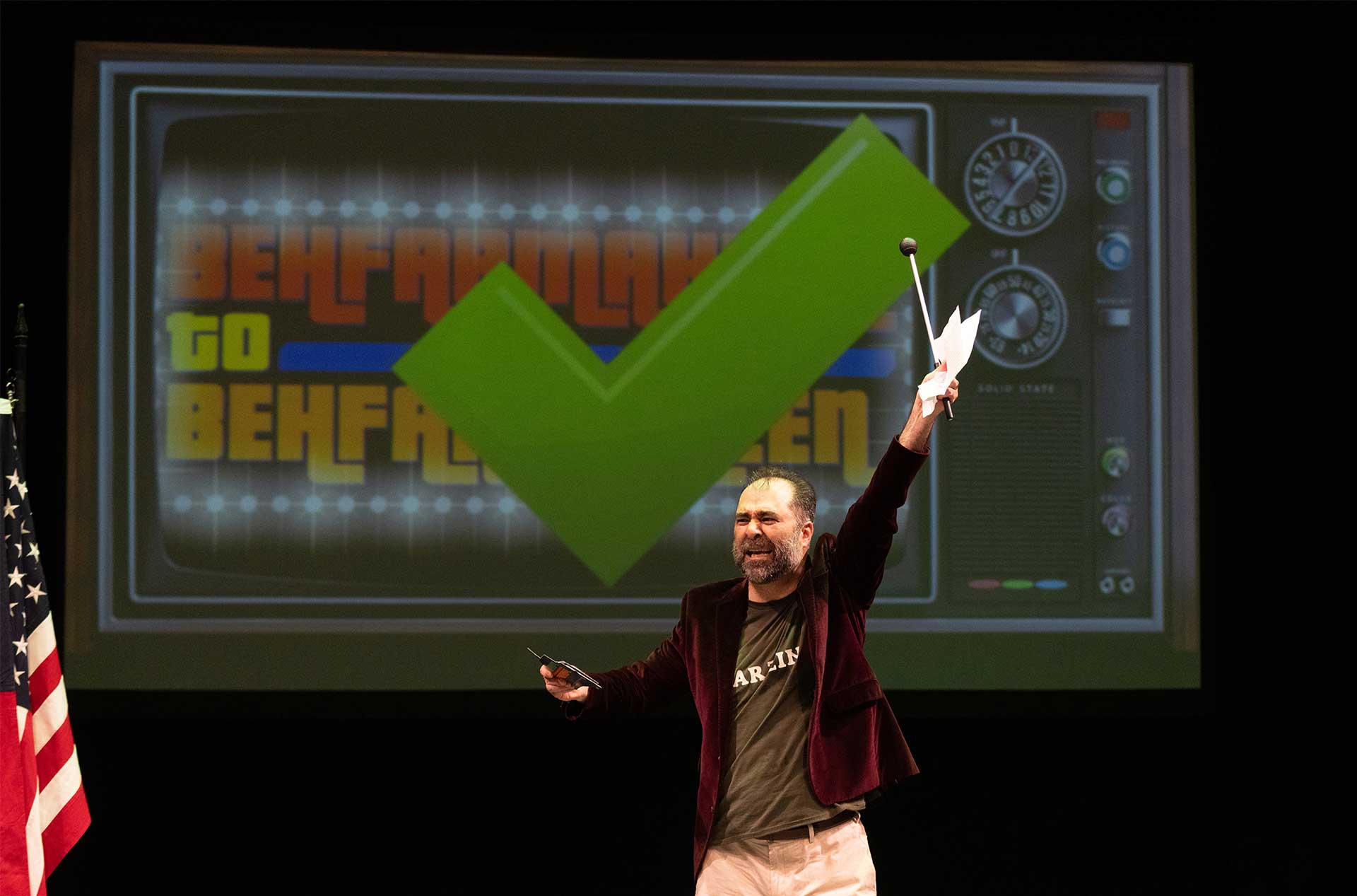 A person triumphantly raises one hand while holding a pointer stick in the other, standing between two flags with a large green checkmark displayed on the screen behind.