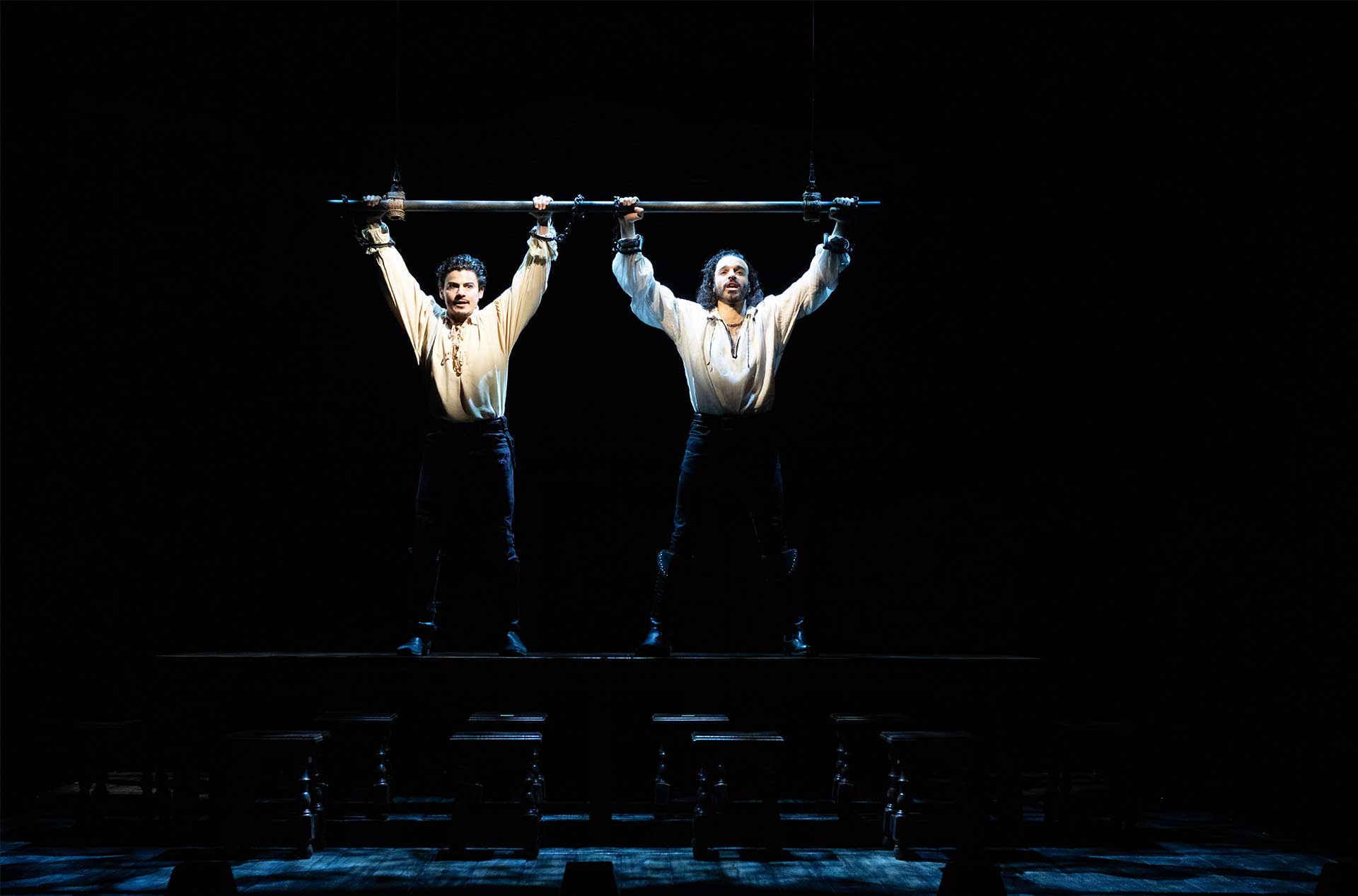 Two people in matching outfits are suspended in the air, each holding onto a horizontal bar with handcuffed hands, illuminated against a dark background.