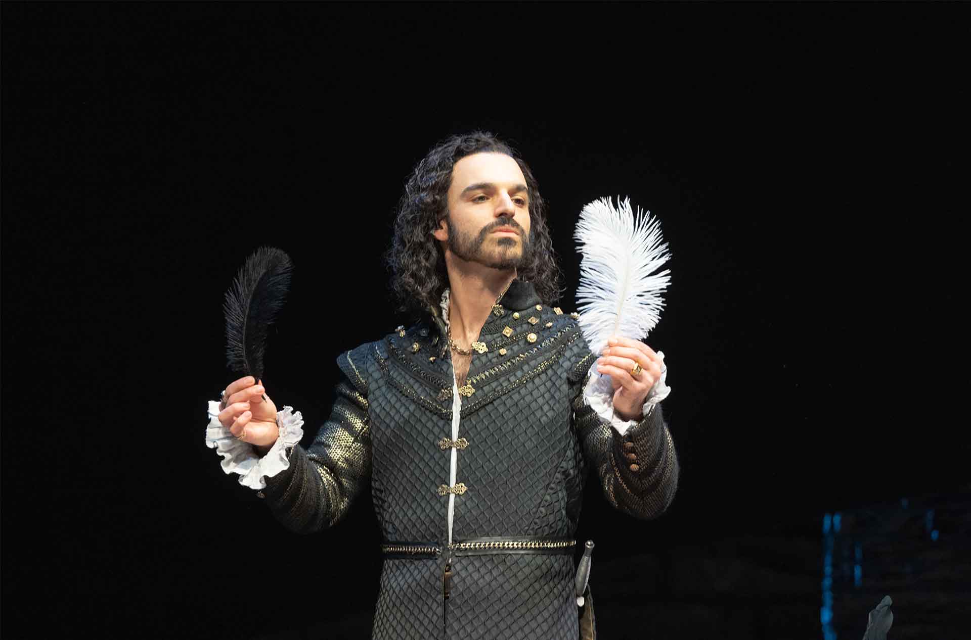 A person with long curly hair and a medieval-style outfit is holding a black feather in one hand and a white feather in the other, looking at the white feather.