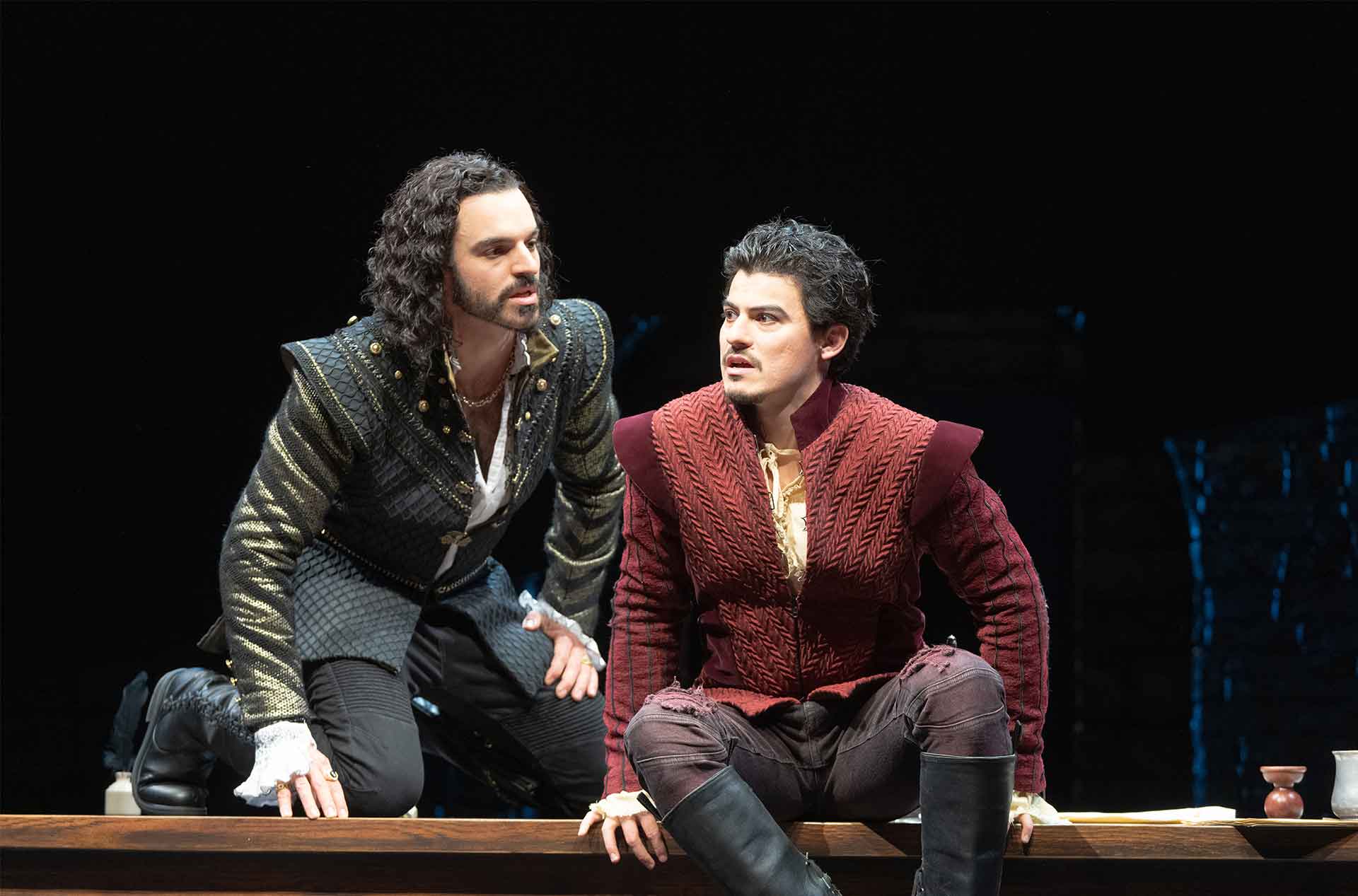 Two people in medieval-style outfits are at a wooden table. The person on the left, with long curly hair and a black outfit, is kneeling and looking at the person on the right. The person on the right, with short dark hair and a red quilted outfit, is sitting and looking into the distance.