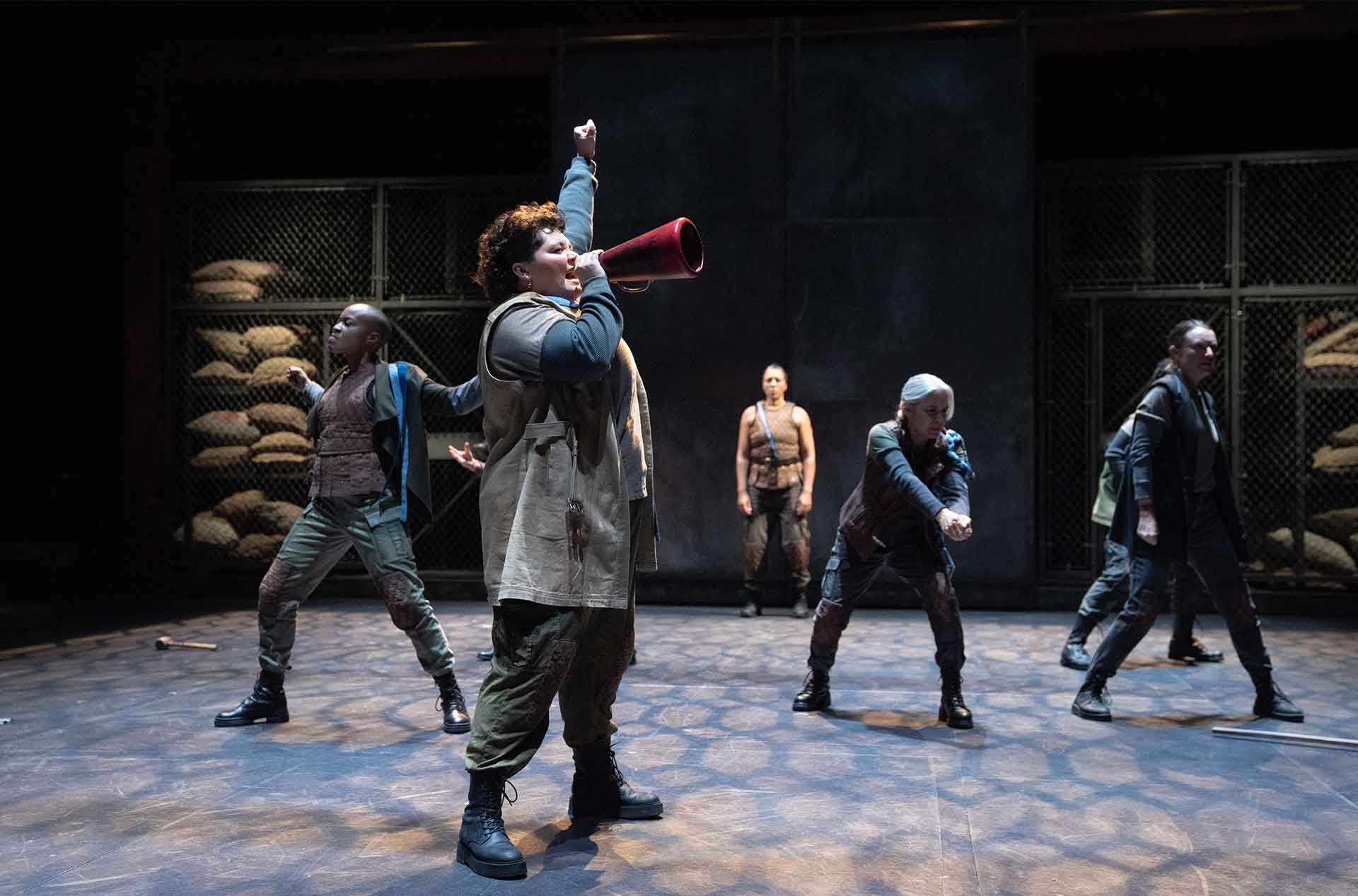 One person energetically leads a group striking dynamic poses on stage while holding a megaphone. The scene set against a backdrop of stacked sacks suggests a rally or call to action.