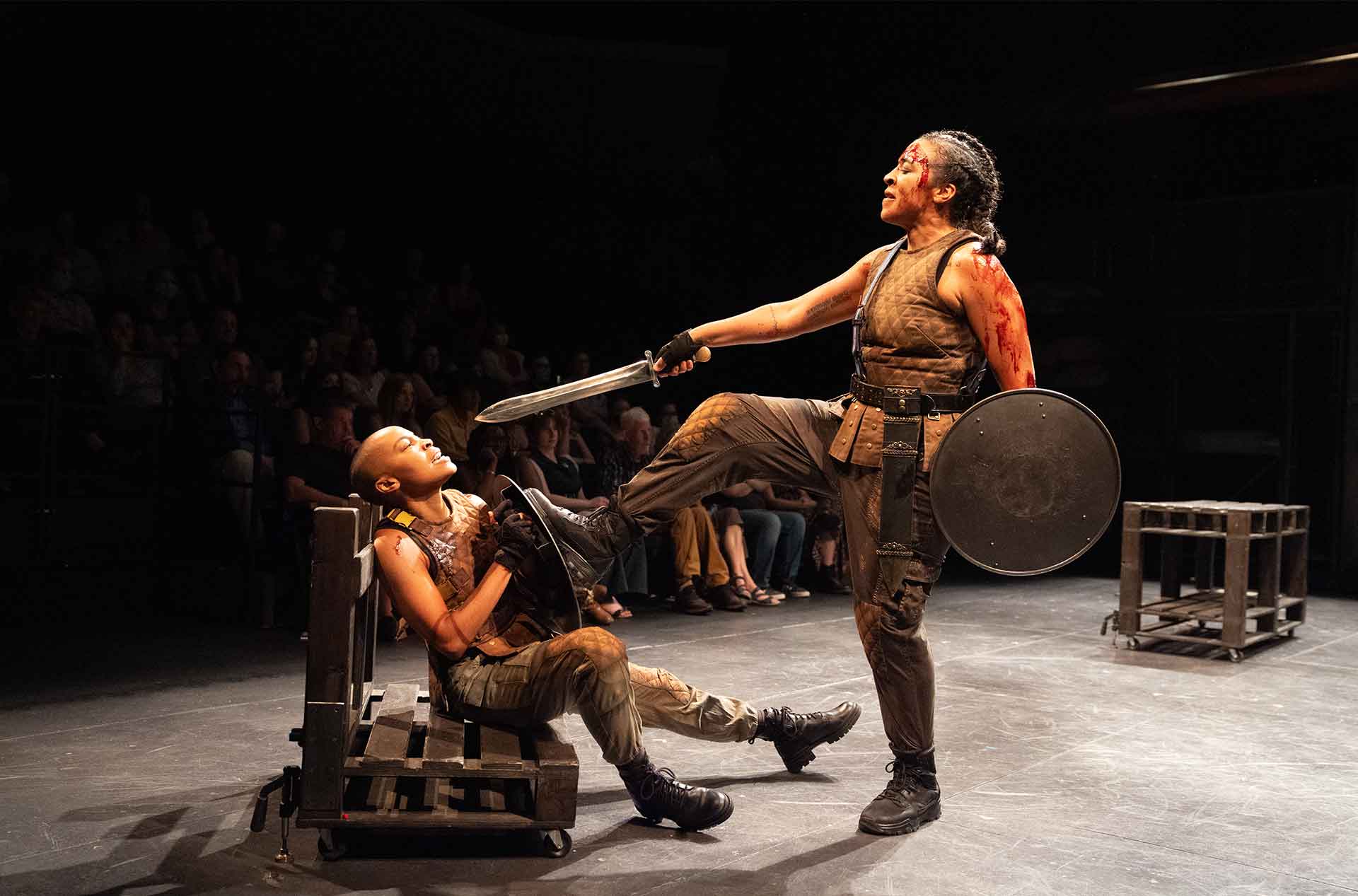 A person clad in a battle-worn costume with a shield and sword stands triumphantly with one foot on another's chest. The seated person holds a shield defensively with an audience visible in the background.