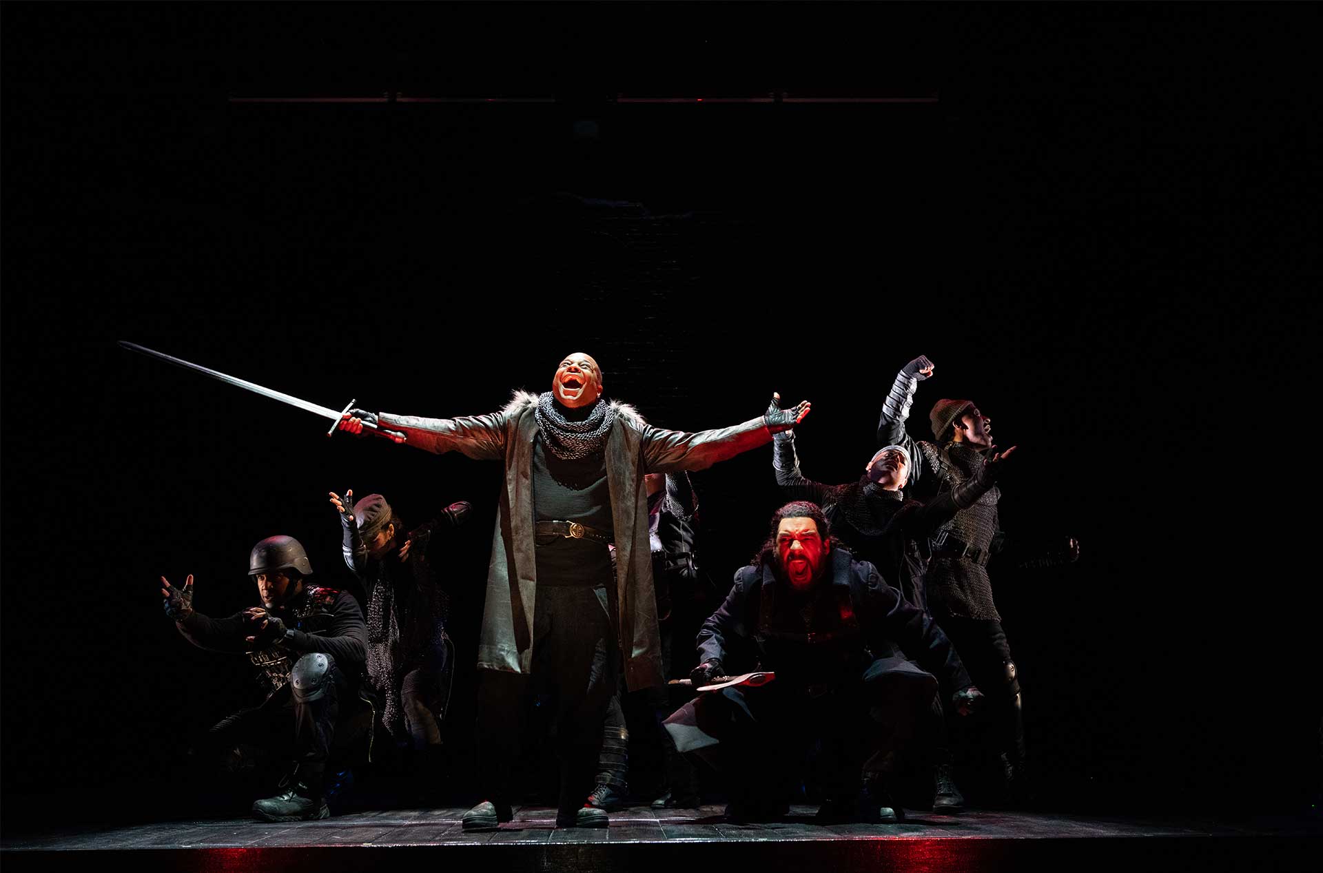 A group of people in dark, medieval-style costumes are on stage. The central figure, standing with arms outstretched and holding a sword, is illuminated and appears to be shouting. The others around them are in various dramatic poses, some crouching and some raising their arms.