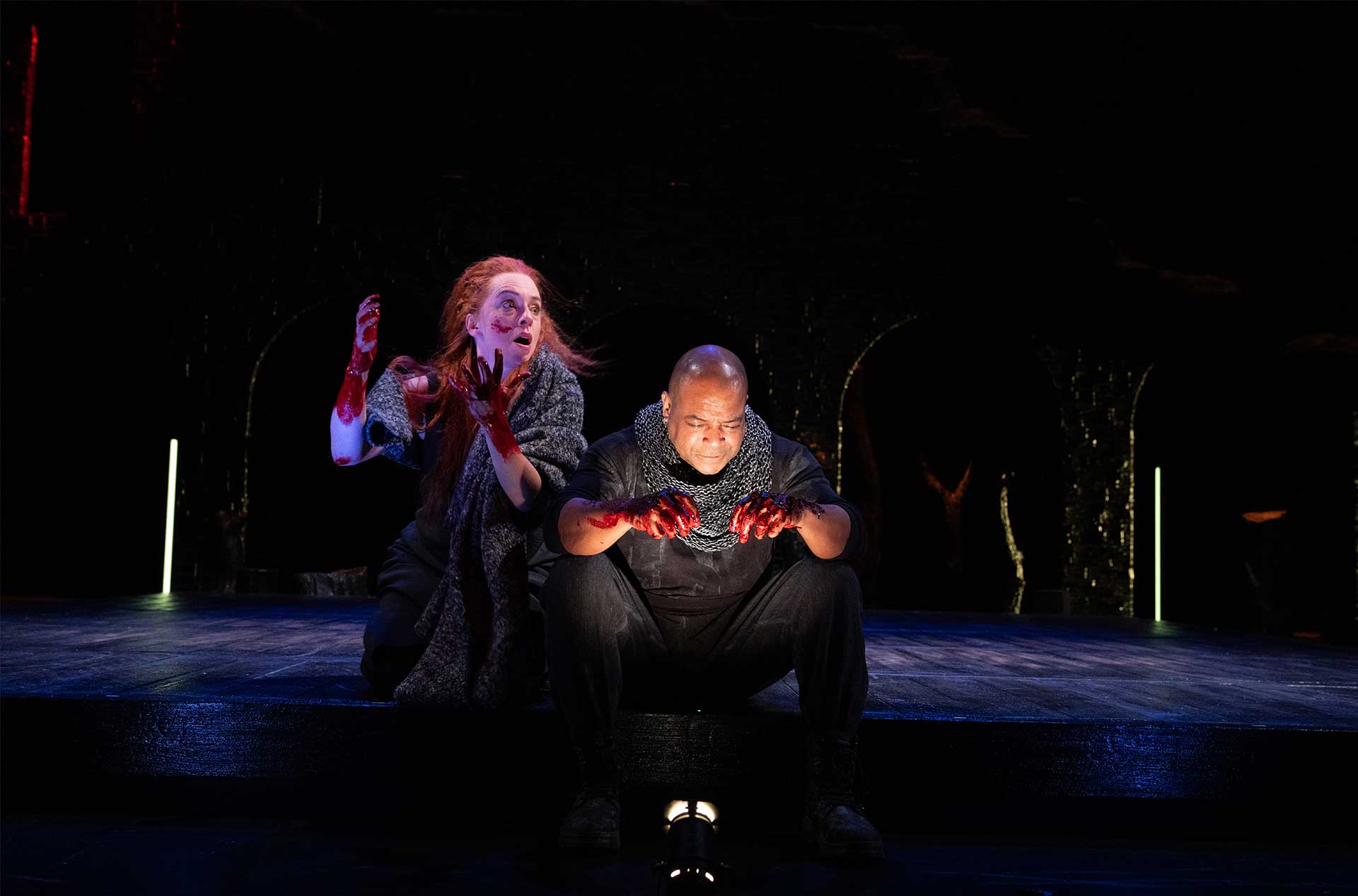 Two people in dark, medieval-style costumes are on stage with blood on their hands. Both are displaying distressed expressions; one person with red hair is kneeling and looking upwards, while the other person is seated, looking down.