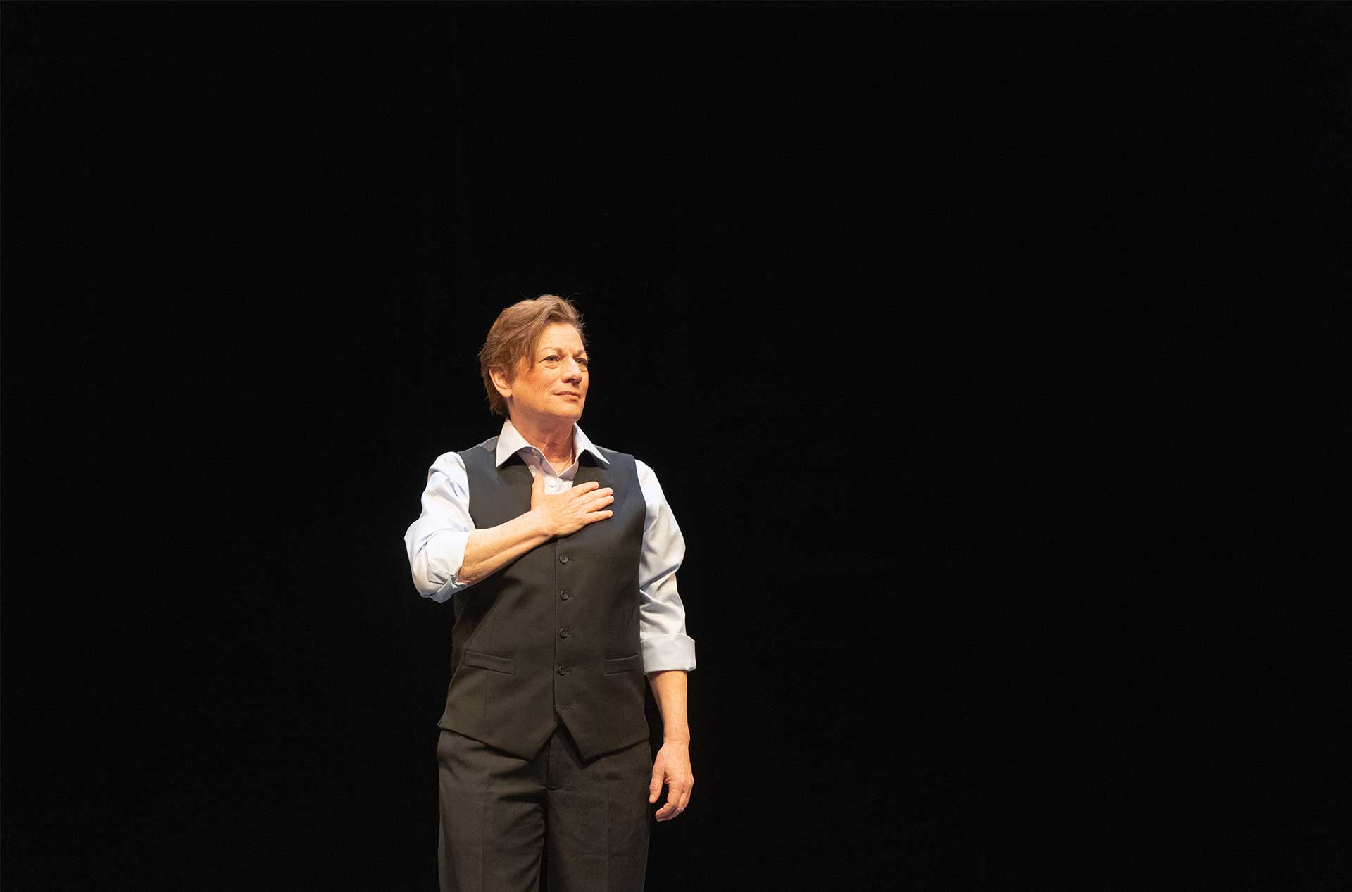 A person standing before a dark background with their hand covering their heart.