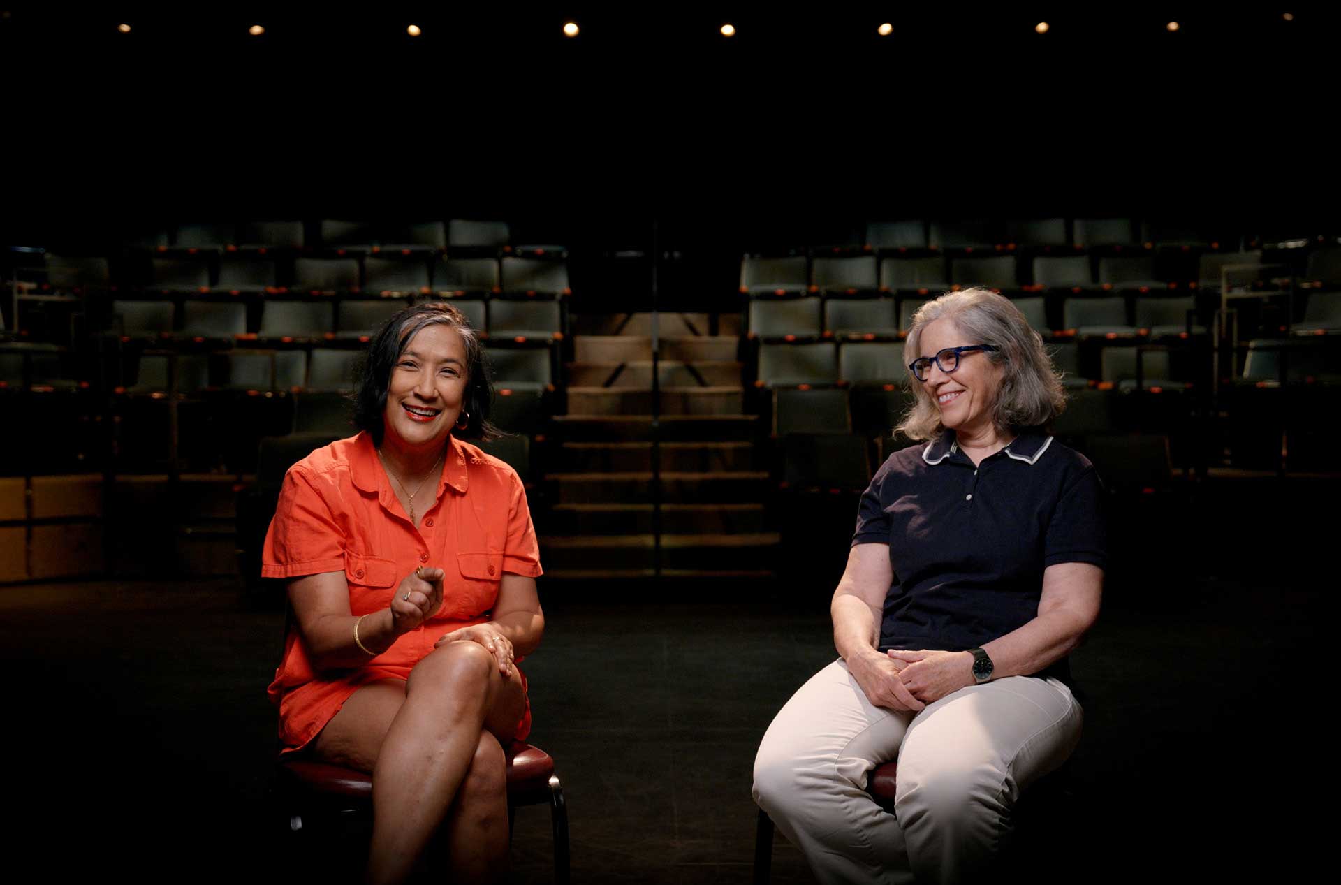 Two people are seated on chairs in a dimly lit theater with empty seats in the background. The person on the left is wearing an orange shirt and shorts, smiling and gesturing with their hand. The person on the right is wearing glasses, a navy blue shirt, and light-colored pants, smiling and looking towards the person on the left.