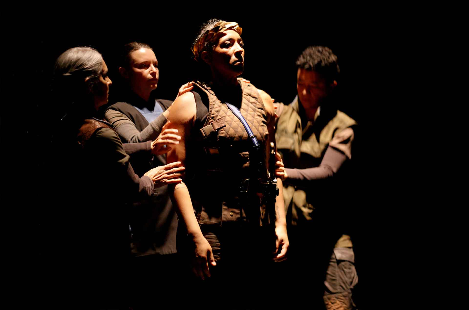 Four people stand in a dimly lit scene. The central figure, dressed in tactical gear and standing upright, is being touched or supported by three others, whose hands rest gently on their shoulders and arms.