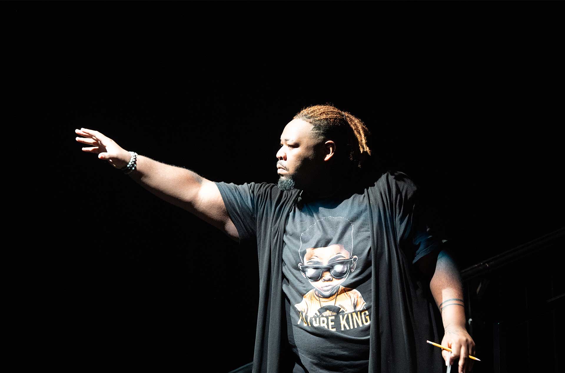 A person stands alone, partially lit, with one arm stretched forward while wearing a shirt that reads “Future King.”