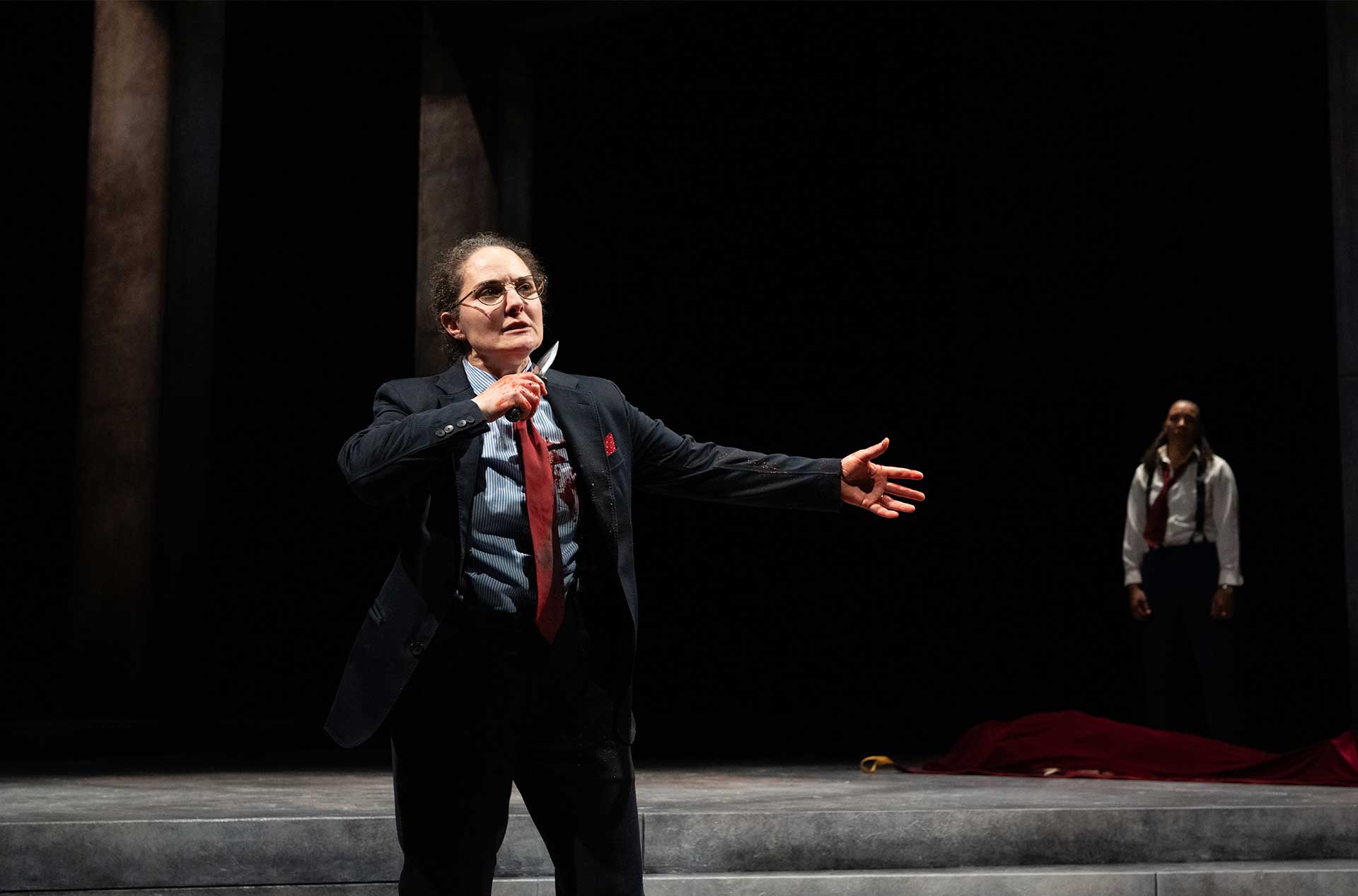 A character in a suit with a loosened red tie holds a dagger to their chest, a bloodstain visible on their shirt, while another figure stands in the distance near a draped red cloth.