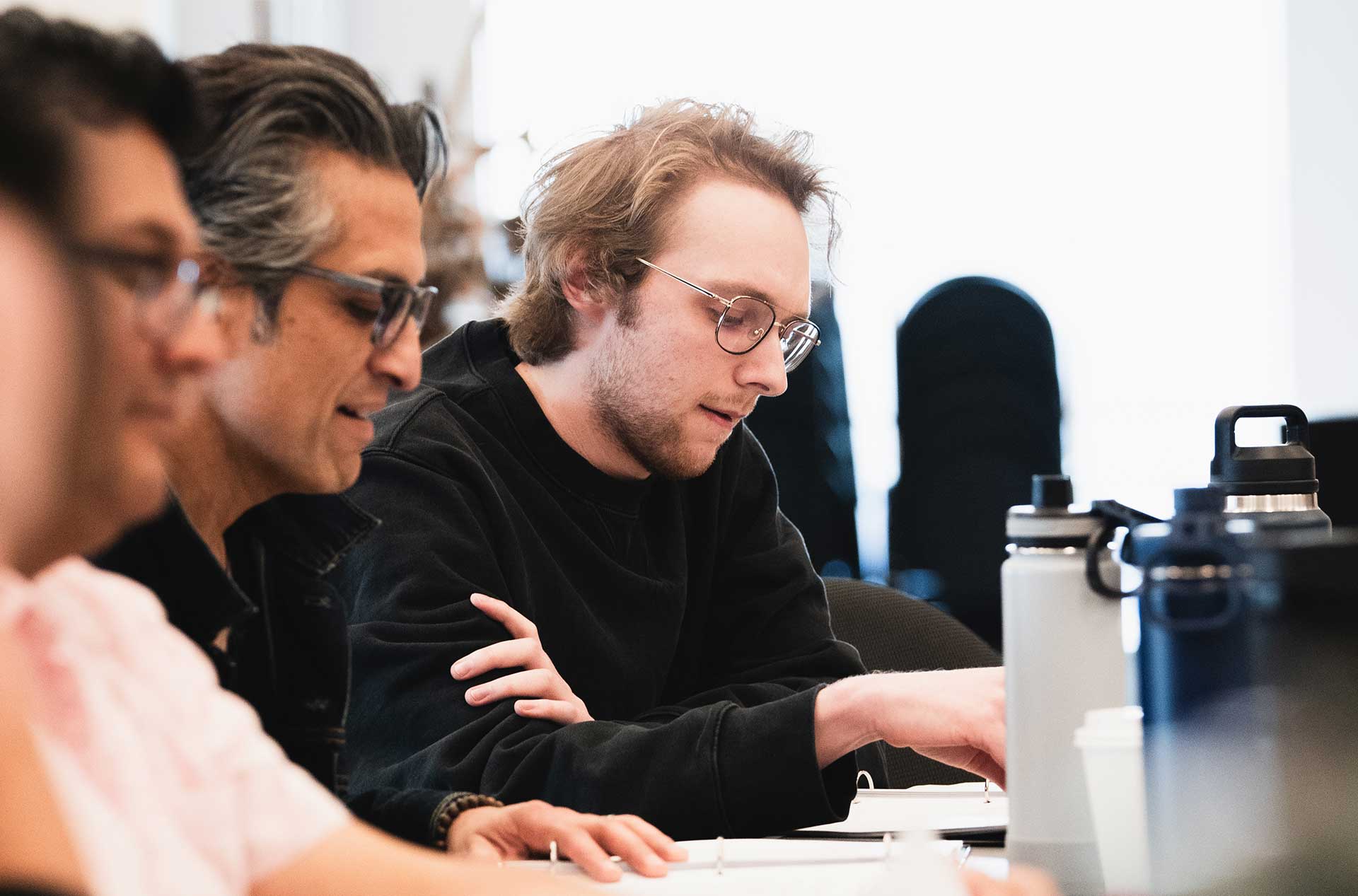 A person in a black sweater looks down at a script, focused. Others nearby are also reading or discussing their materials, with large water bottles on the table.