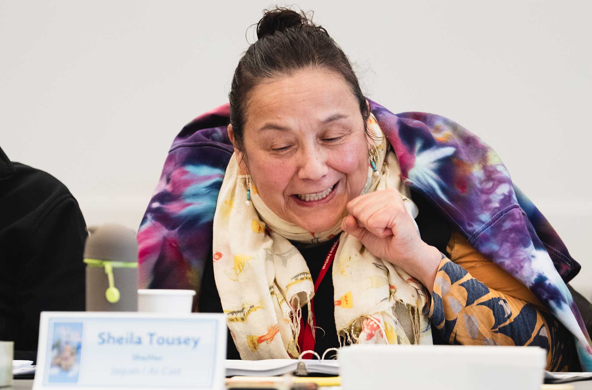 A person wearing a vibrant tie-dye hoodie and a patterned scarf smiles while looking down at a script. 