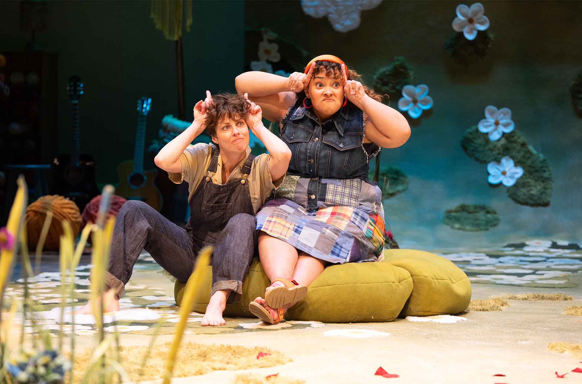 Two seated performers on stage make playful antler gestures with their hands, sitting on soft green cushions amid a whimsical set dotted with guitars and giant felt flowers.