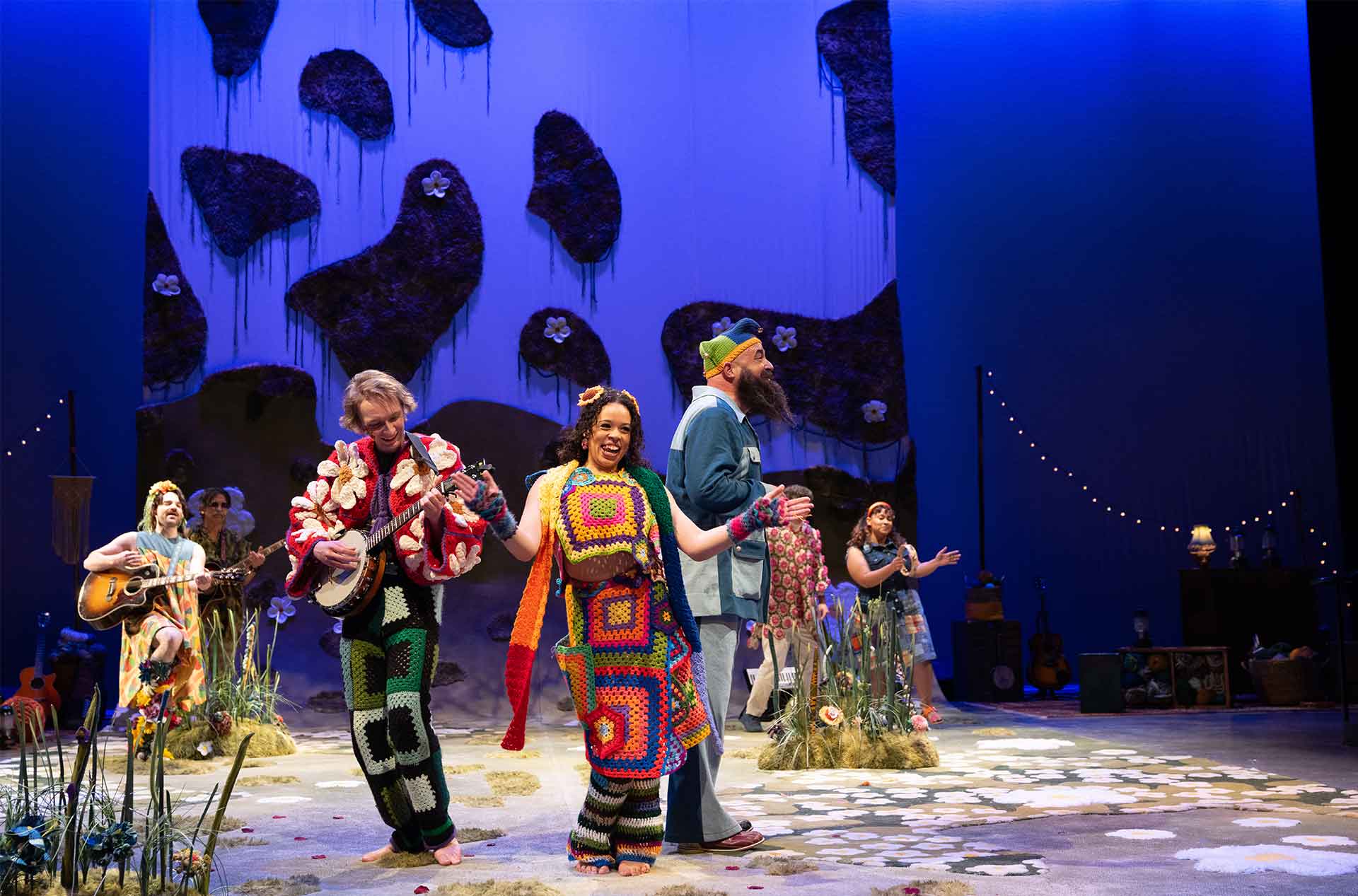 Performers in colorful crocheted and patchwork costumes sing and play instruments on a whimsical stage decorated with large floral patterns and grassy textures.