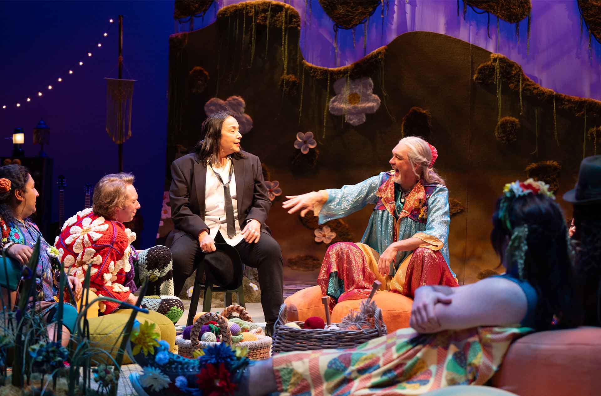 Two seated performers talk animatedly; one in a floral cloak gestures while the other, dressed in a black suit and loose tie, listens in an intimate, casual setting onstage.