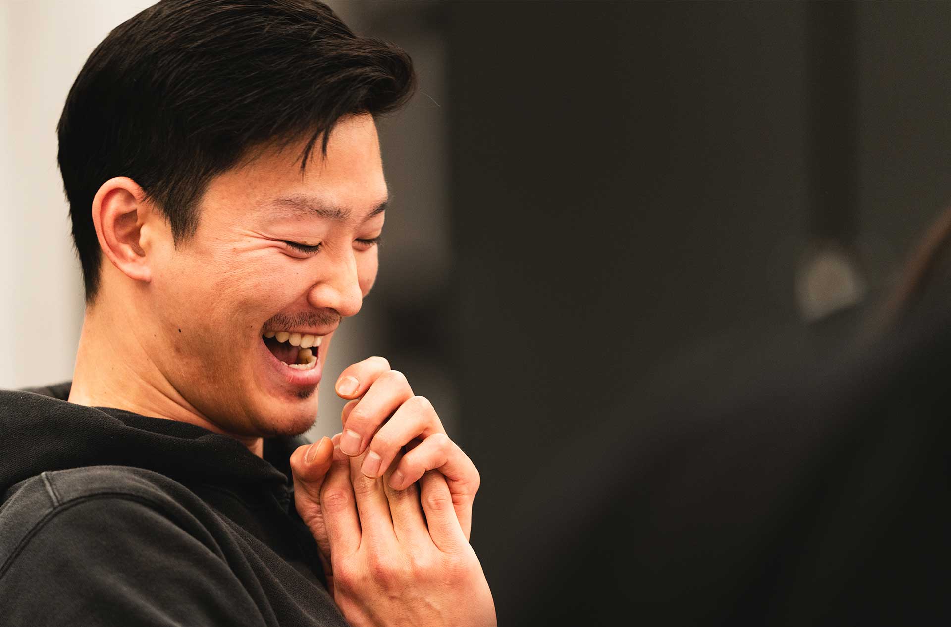 A person laughs openly, hands clasped together, while engaging in conversation during the rehearsal. The background is dark and out of focus, highlighting the expression.