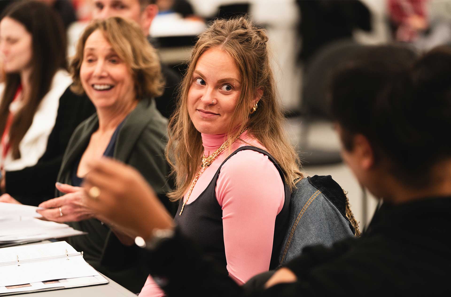 A person in a pink shirt layered under a black top listens intently while seated in a group discussion. Other individuals in the background are engaged, with some smiling or speaking.