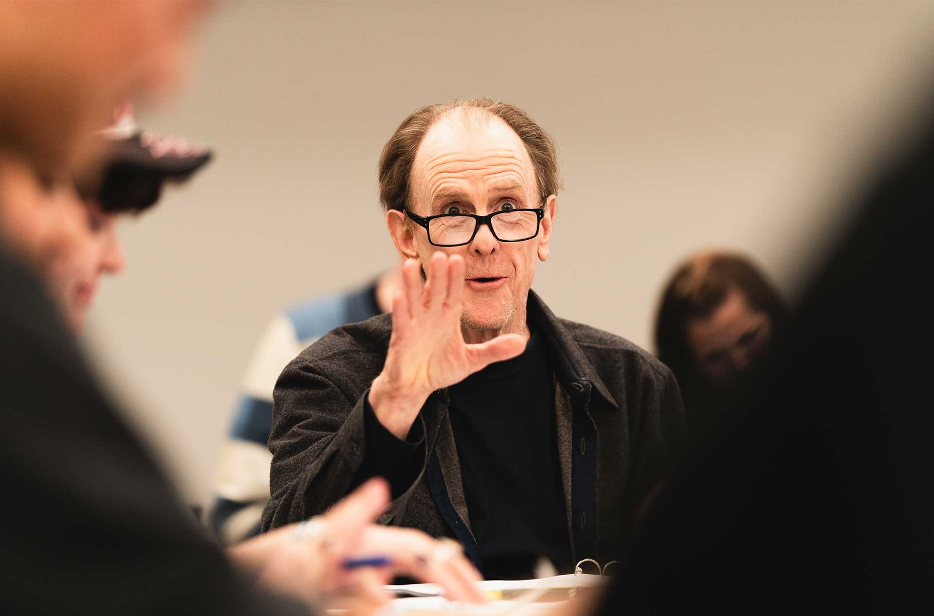 A person with glasses gestures mid-discussion during the rehearsal, their expression animated. Others in the background are listening and taking notes.