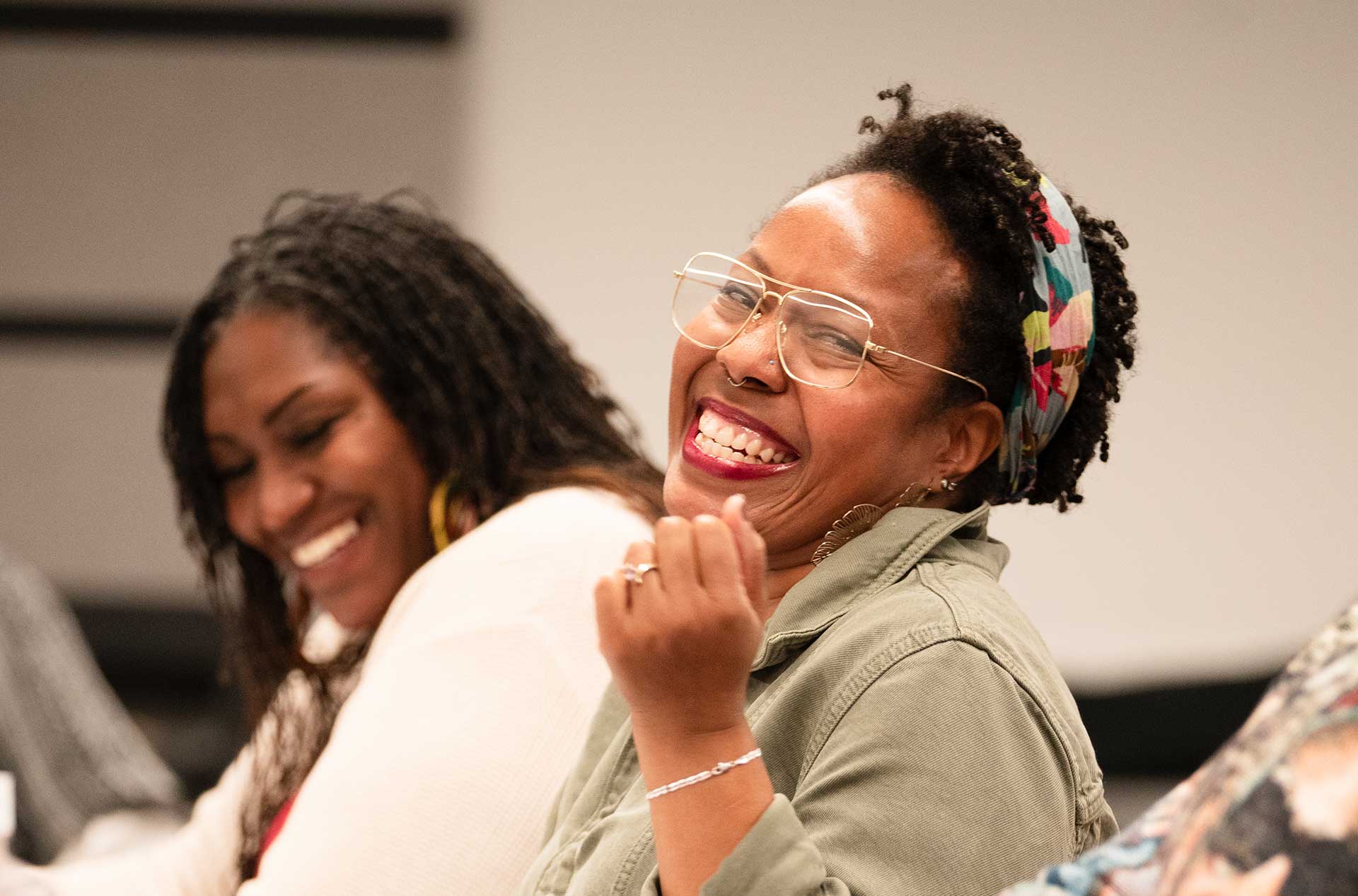 A person wearing glasses and a green jacket laughs while sitting at a table. Another person in the background, also smiling, appears to be engaged in the conversation.