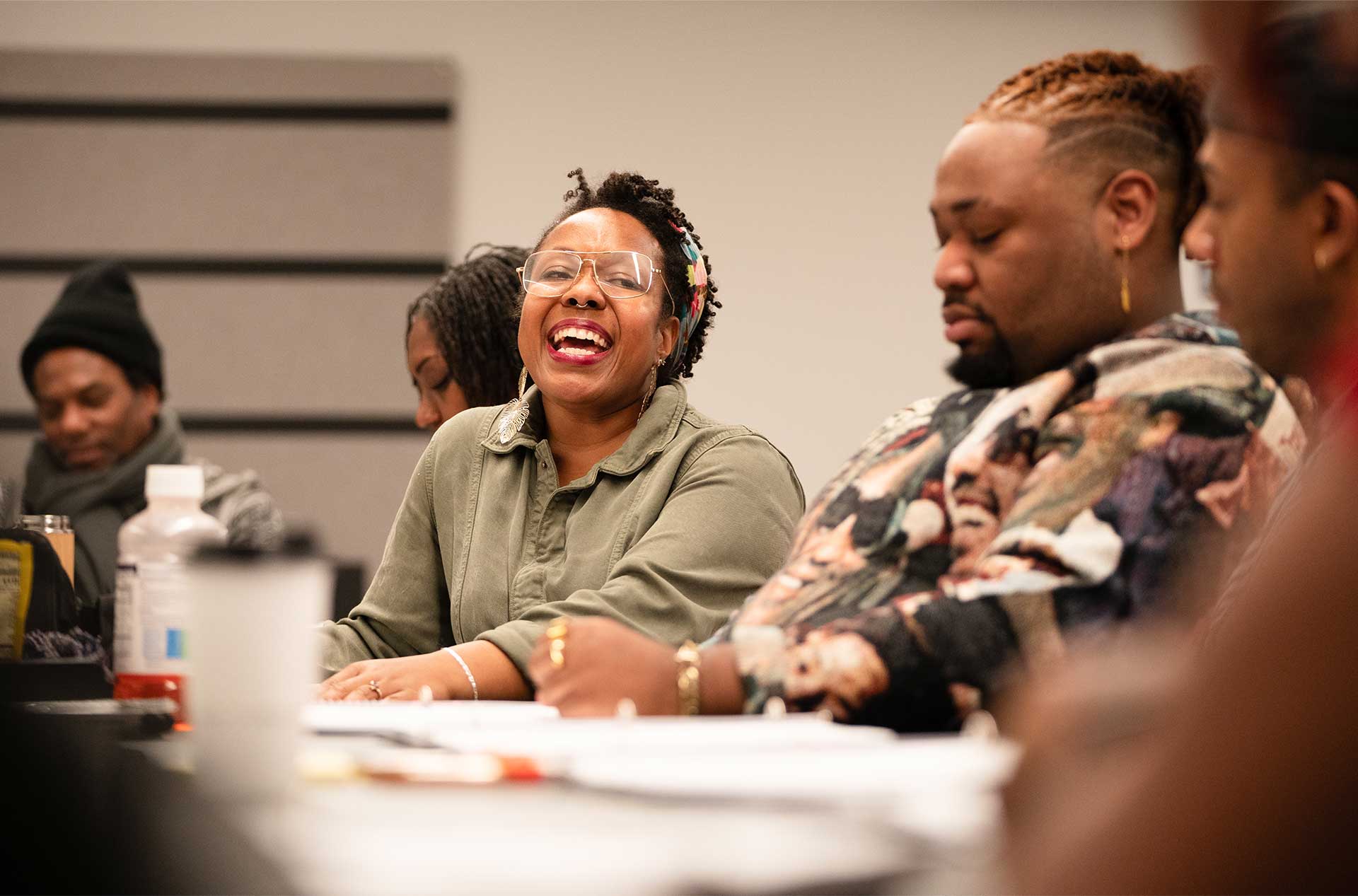 A lively discussion at a table, where a person in a green jacket and glasses laughs with an open mouth. Others around them appear to be listening or reacting.
