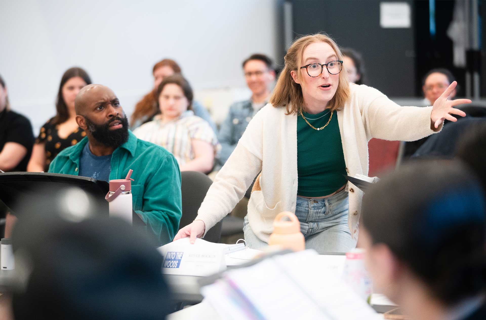 A person gestures passionately during a rehearsal while standing, with another seated person looking on. 