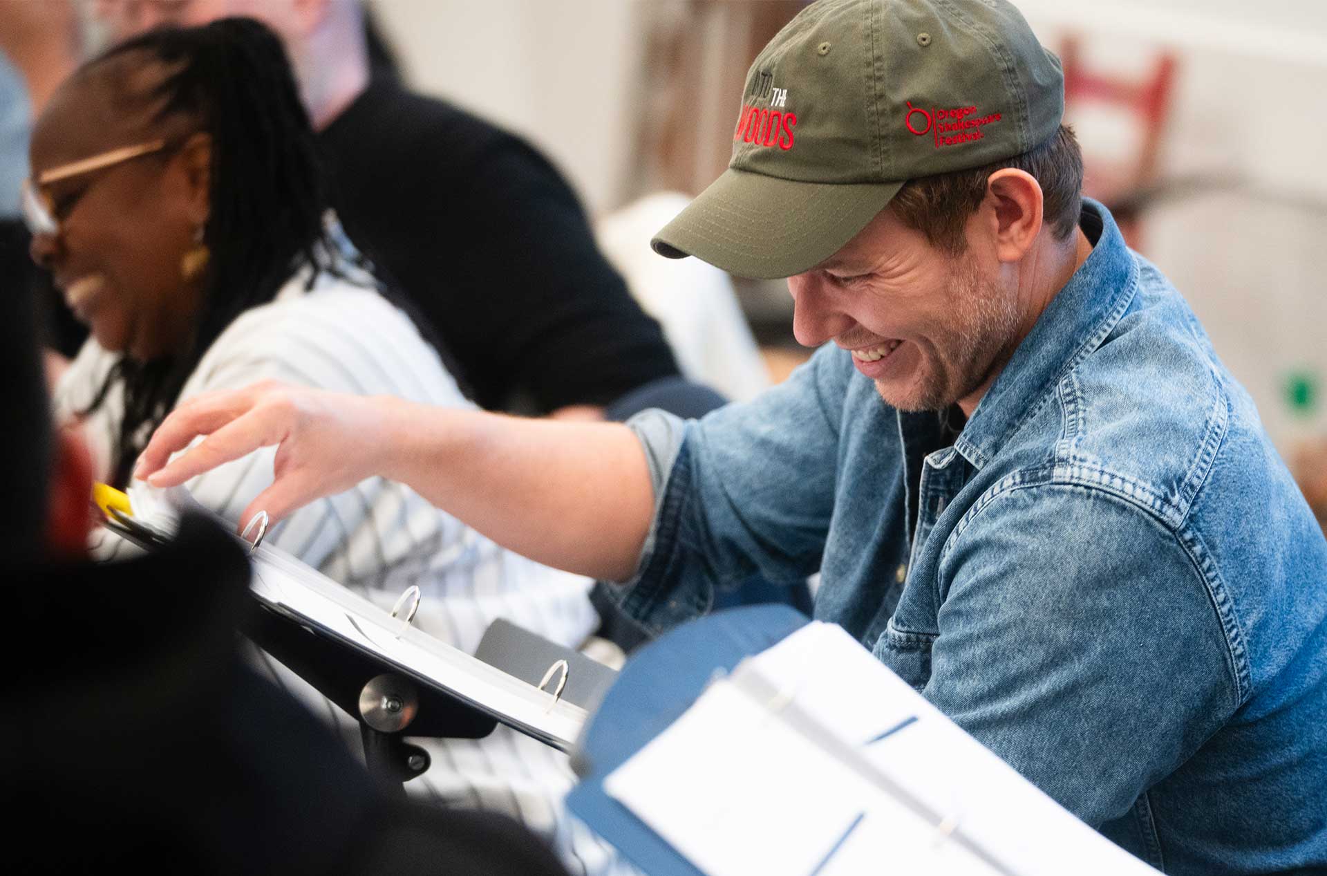 A smiling person wearing a green "Into the Woods" cap and denim shirt laughs during a table read, surrounded by others holding scripts.