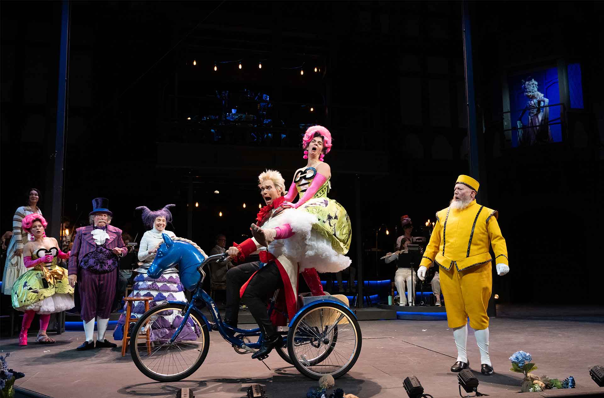 A theatrical moment on stage as a performer in bright red guides a tricycle with a blue horse head, while another in a poufy yellow and green dress rides in back; onlookers in colorful costumes watch.