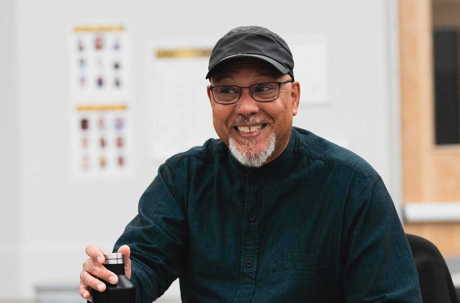 A person with glasses and a black cap sits at a table, smiling broadly while holding a water bottle.