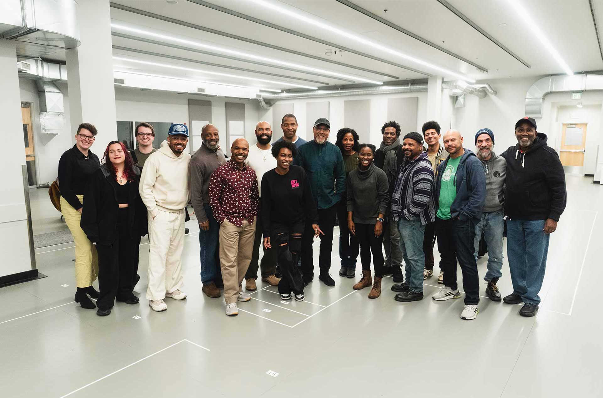 A diverse group of people standing together in a spacious room, smiling for a group photo. Everyone appears relaxed and happy, with some people casually posing with hands in their pockets.
