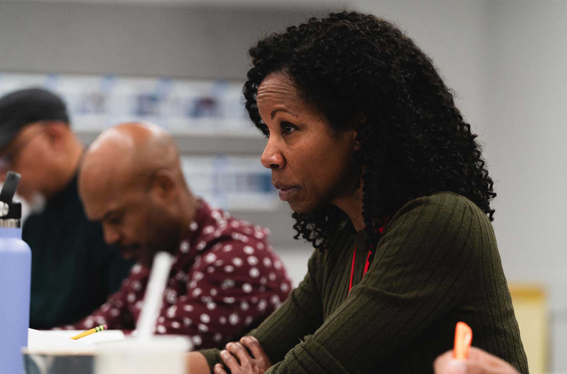 A person with curly hair, wearing a green sweater, listens attentively during a rehearsal.