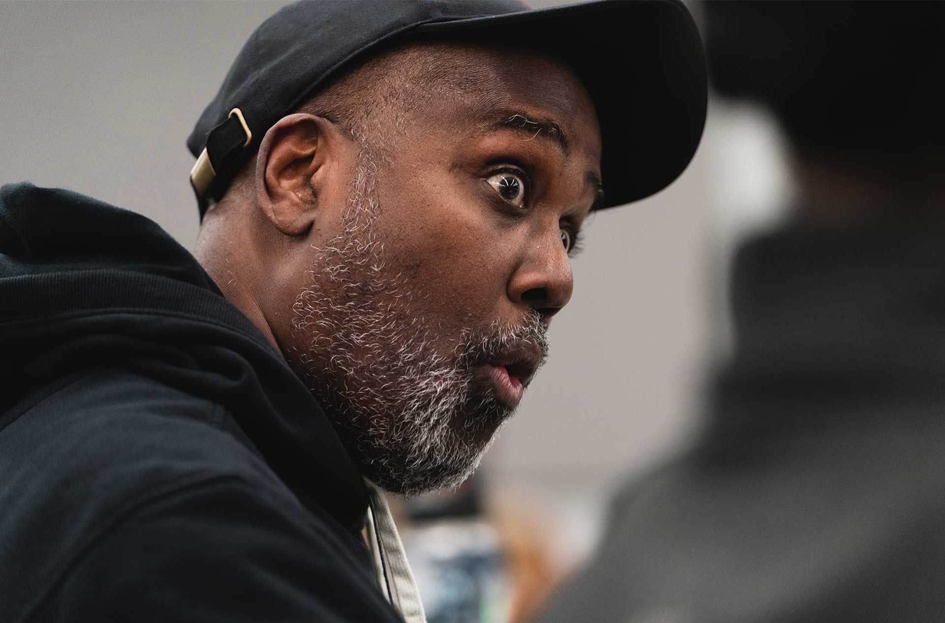 A person with an expressive look, wearing a black hoodie and cap, appears to be mid-conversation.