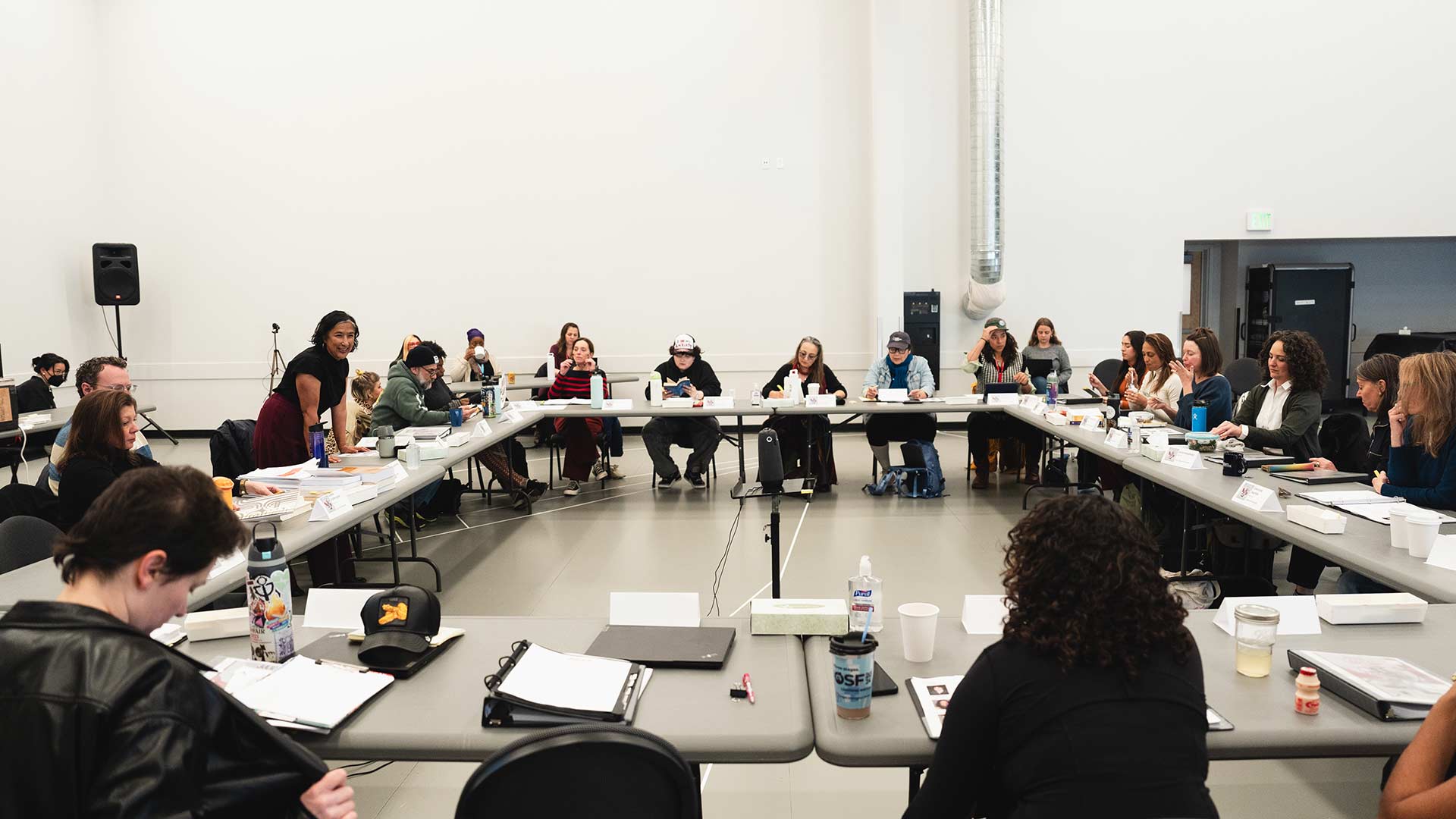 A play rehearsal in progress, with a large group of individuals sitting around rectangular tables arranged in a square. Scripts, notebooks, and drinks are scattered across the tables.