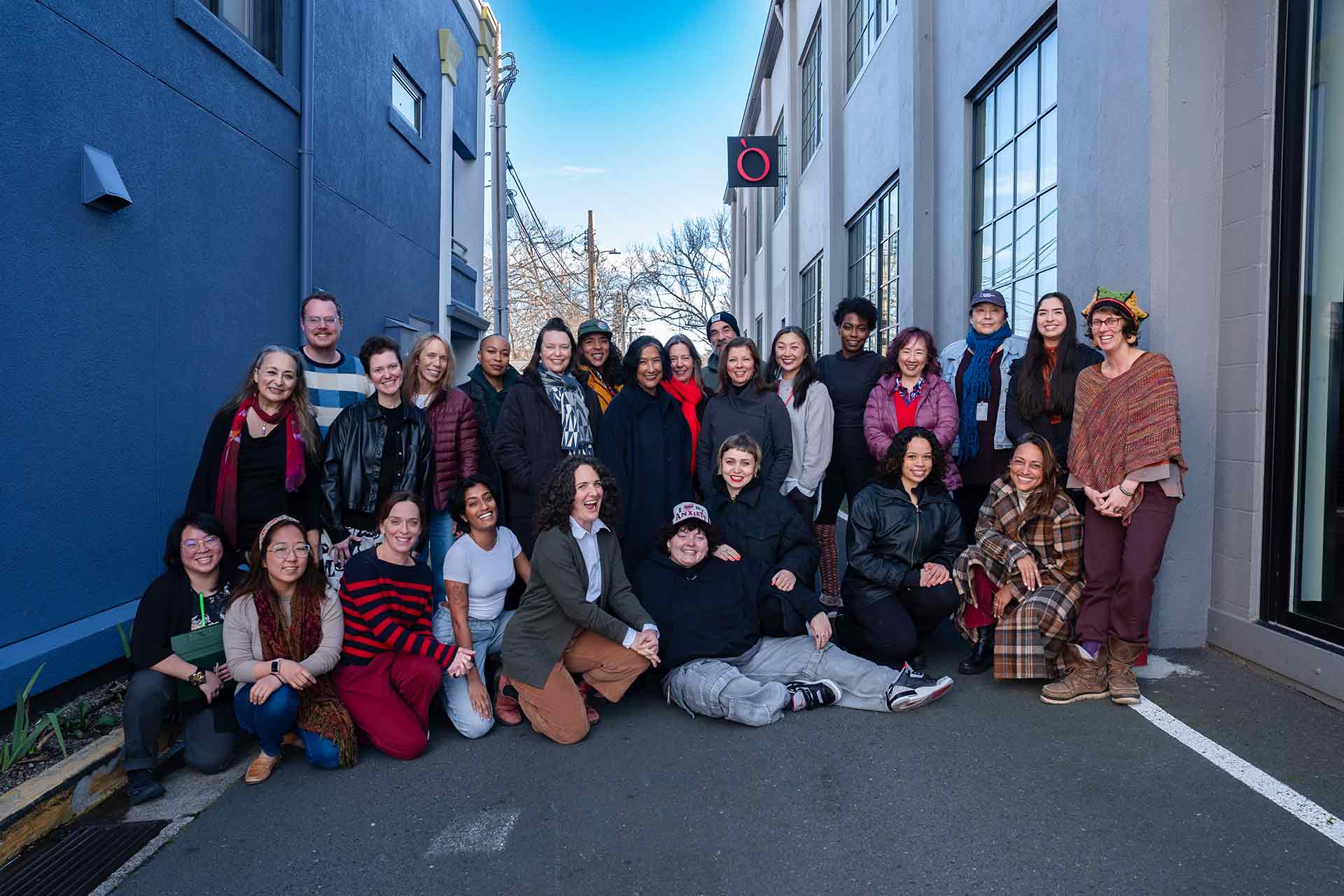 The cast and creative team of a play pose for a group photo outside a building with blue and gray walls. Some are standing, while others are kneeling or sitting in the front row.