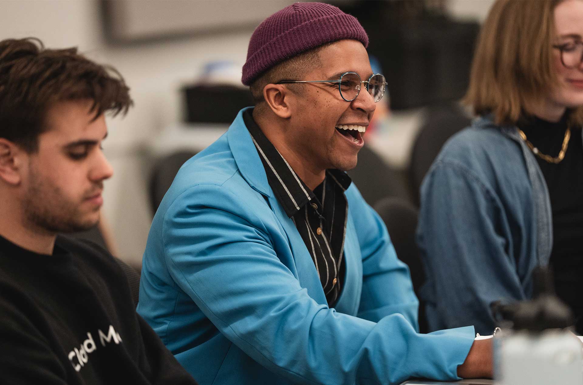 A person in a bright blue blazer and purple beanie laughs during a rehearsal, seated between others reading scripts.