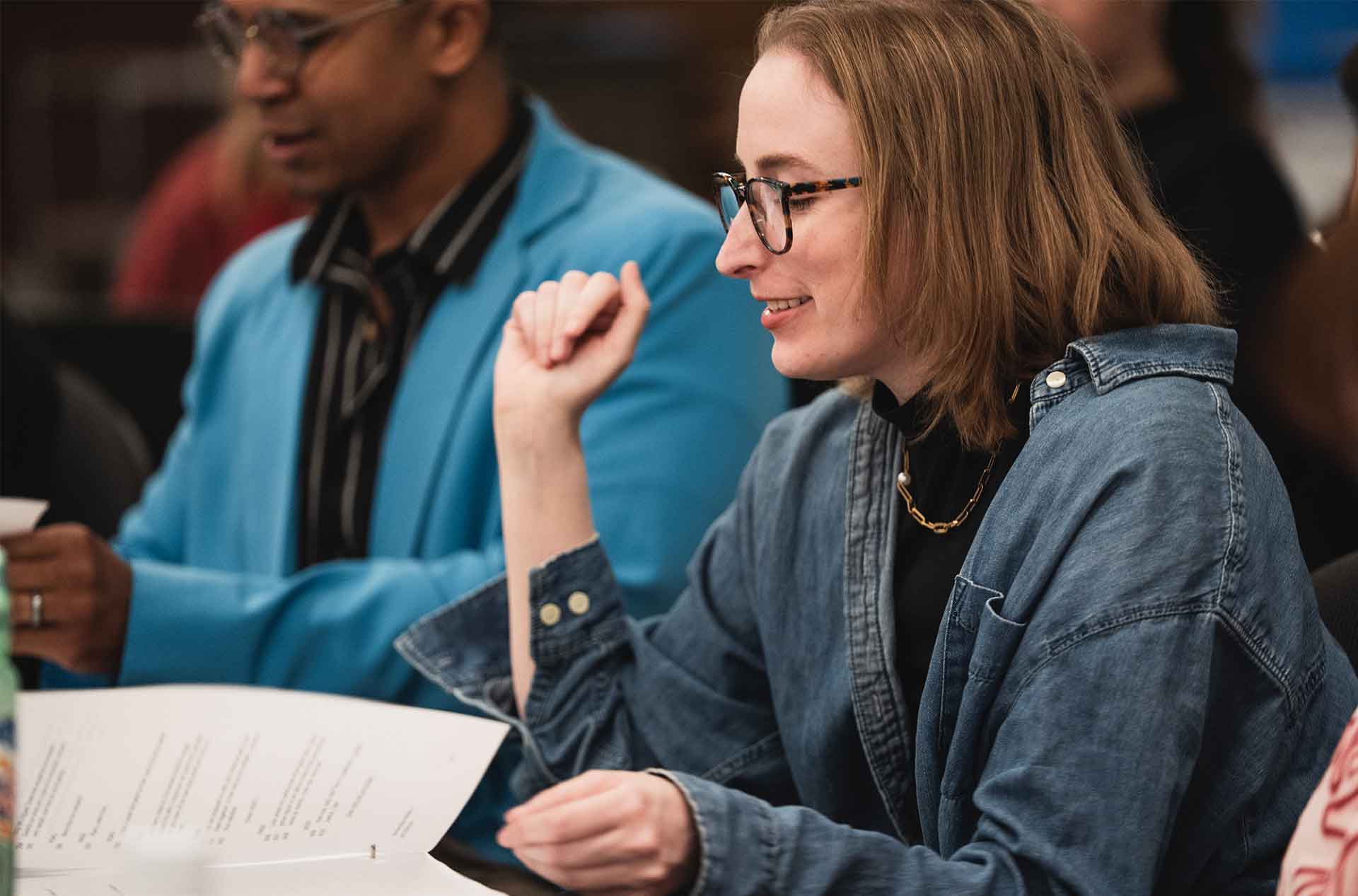 Person wearing glasses and a denim shirt smiles while engaged in conversation, holding a script or printed material during what appears to be a table read or group discussion. Another individual in a bright blue blazer sits nearby, also holding papers.